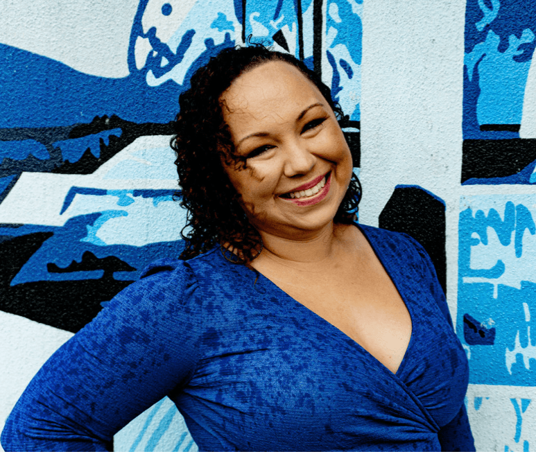 Smiling woman with curly hair in a blue dress standing in front of a blue and white mural.