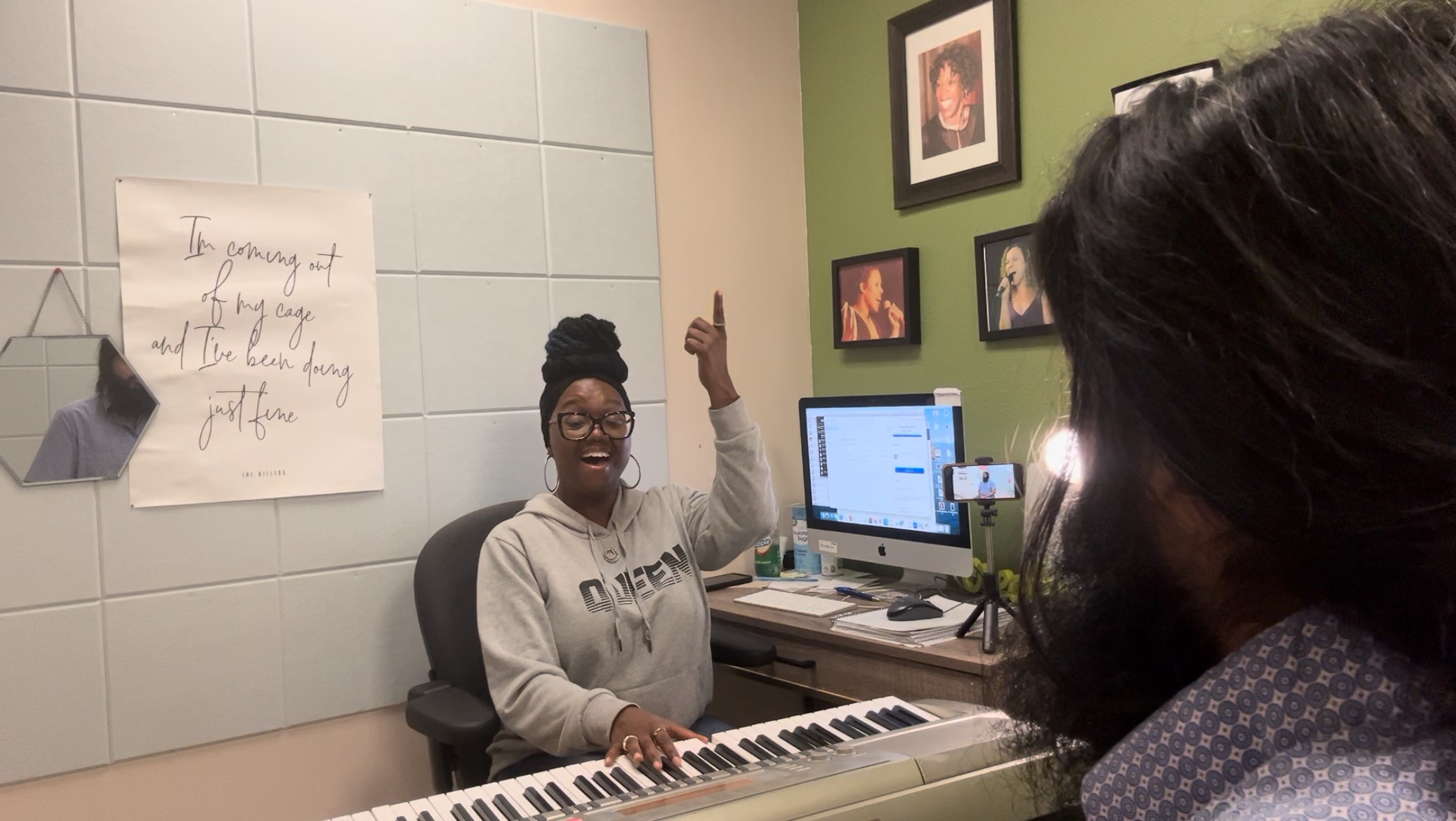 Two women in a room with a computer and keyboard, one smiling and raising her hand, the other facing away.