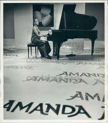 Woman playing grand piano in a room with artwork on the wall, floor has the name 'AMANDA' repeated multiple times.