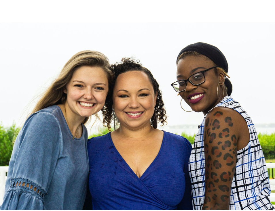 Three women smiling outdoors against a white background, one with long blonde hair, one with curly dark hair, and one with glasses and braids.