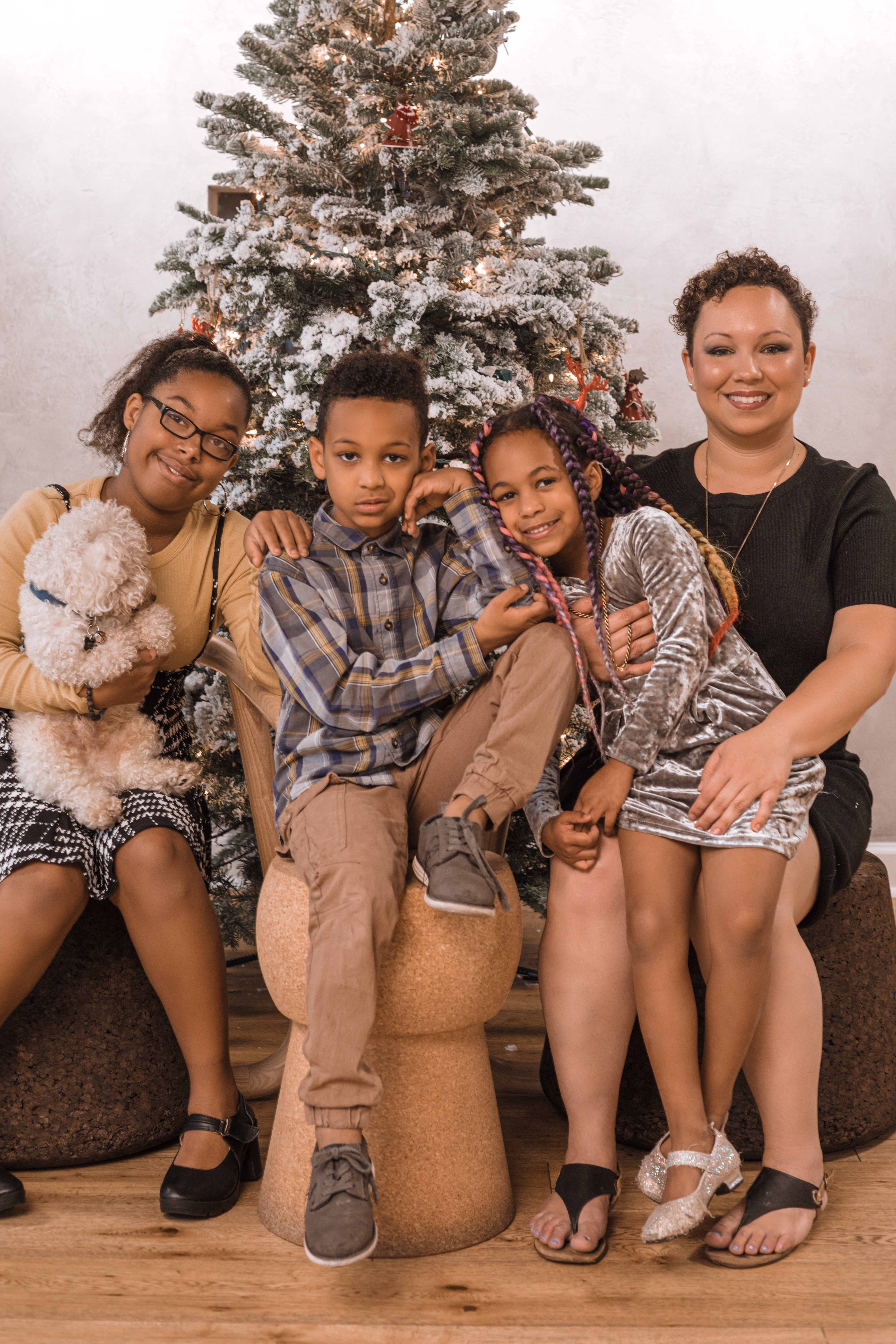 Family of four with a woman and three children sitting in front of a decorated Christmas tree, smiling, holding a dog.