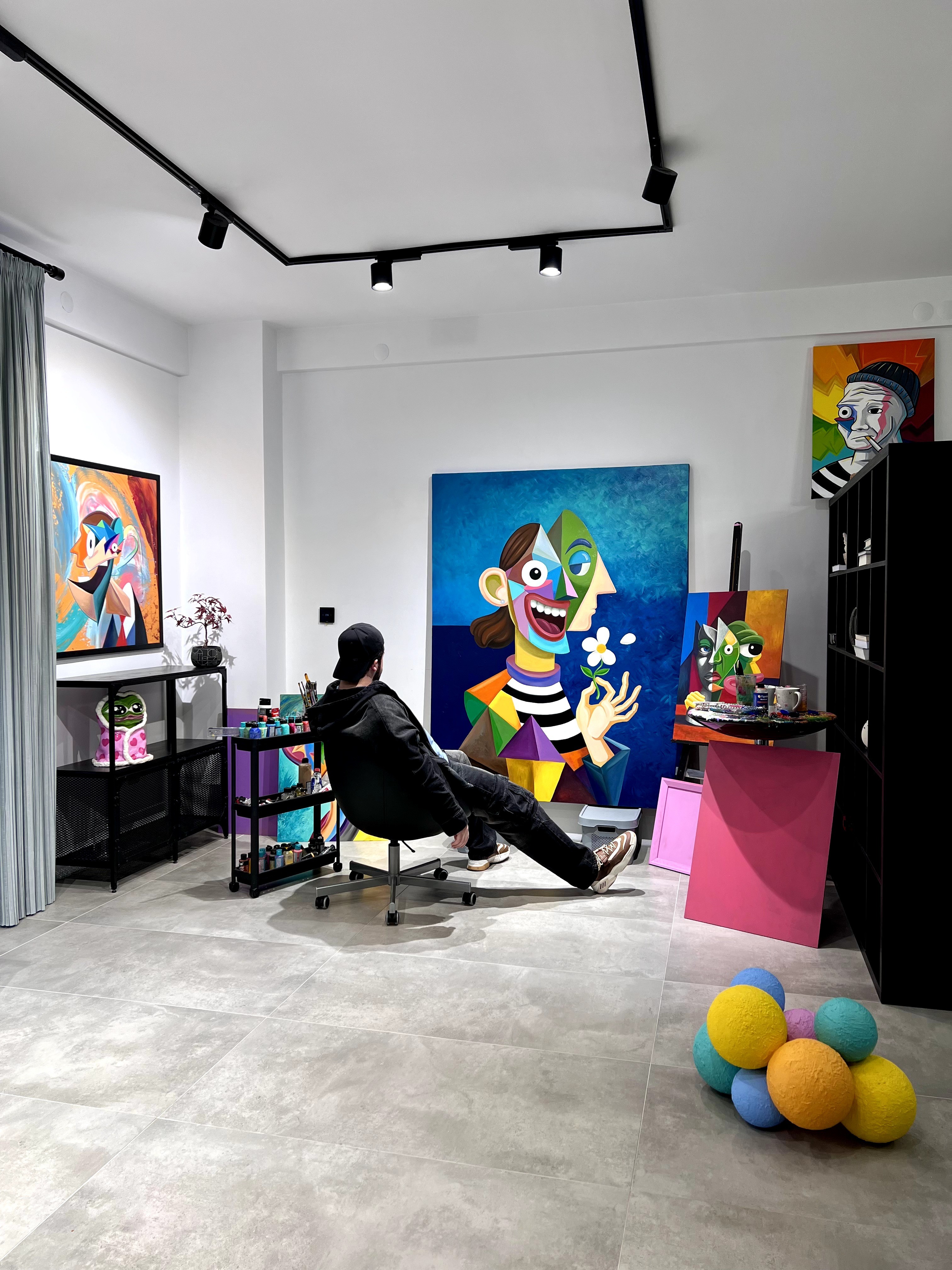 Person sitting in an art gallery surrounded by colorful paintings and balloons on the floor.