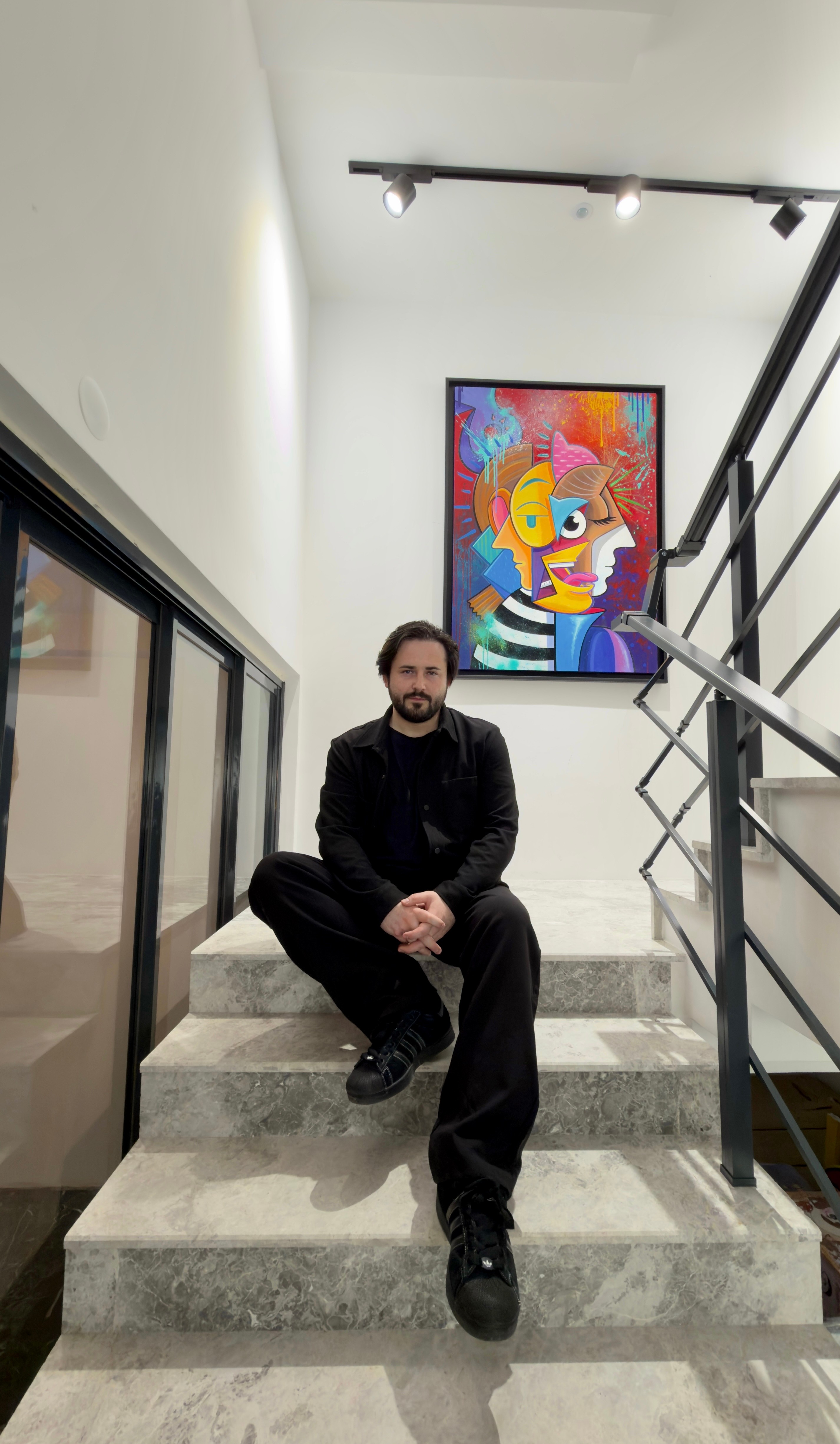 Man sitting on stairs in front of colorful abstract artwork, with railing and window on the side.