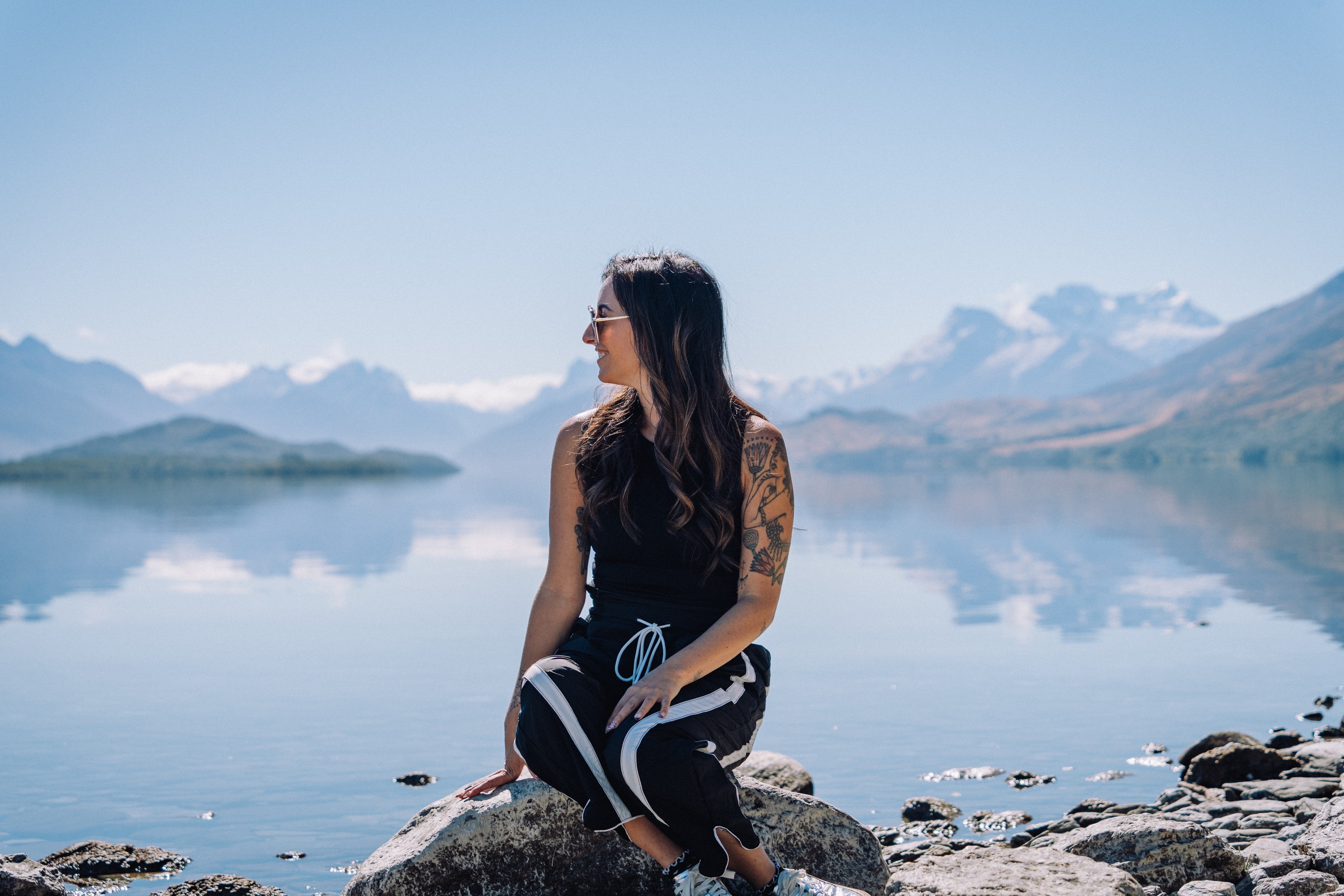 Woman sitting on rocks by a lake with mountains in the background, facing sideways, wearing sunglasses and casual clothing.