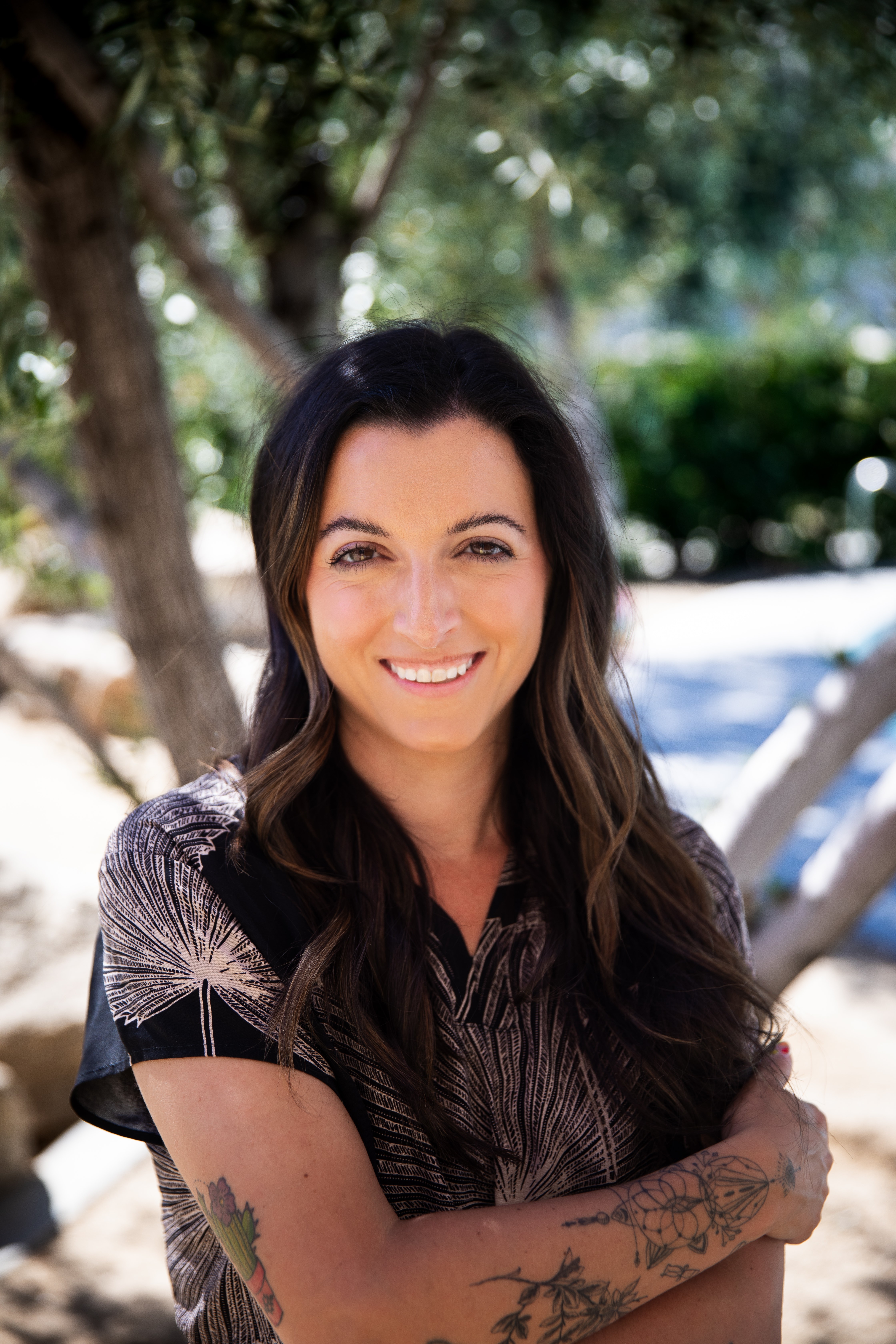 Woman with long dark hair smiling outdoors, arms crossed, trees and sunlight in background.