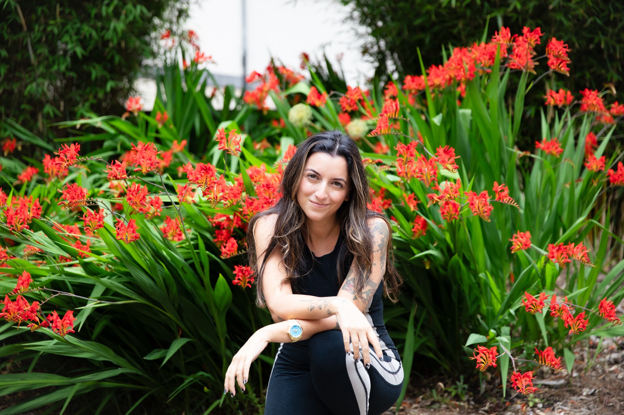 Young woman with long dark hair sitting among red flowers and green foliage outdoors, smiling at the camera.