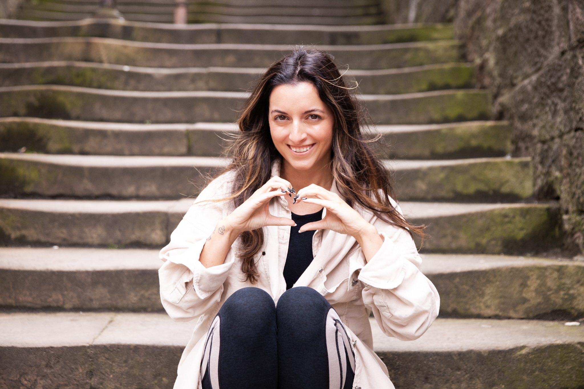 Young woman with long dark hair smiling, sitting on stone steps outdoors, making a heart shape with her hands.