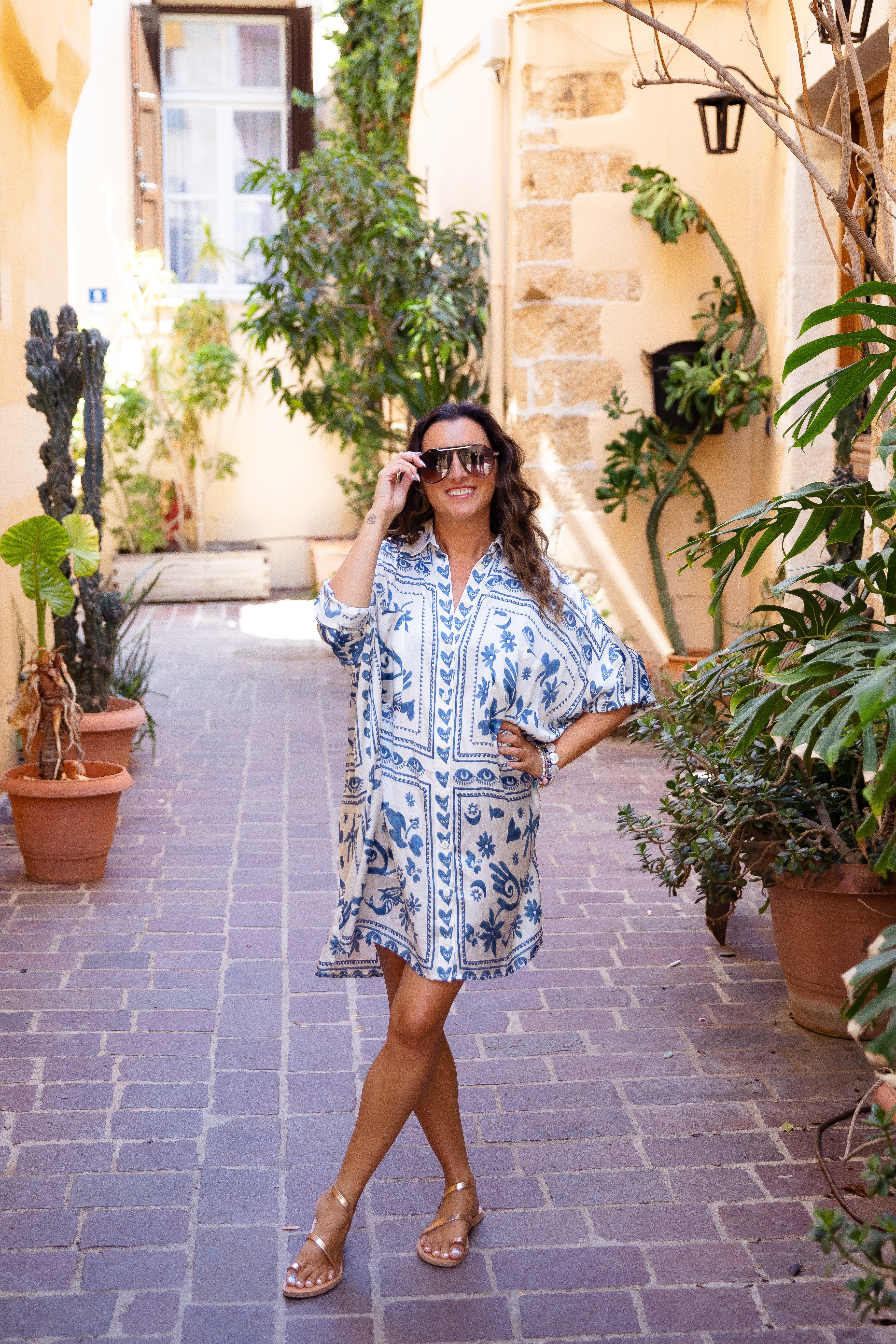 Woman in patterned dress and sunglasses standing on a cobblestone street surrounded by potted plants and buildings.