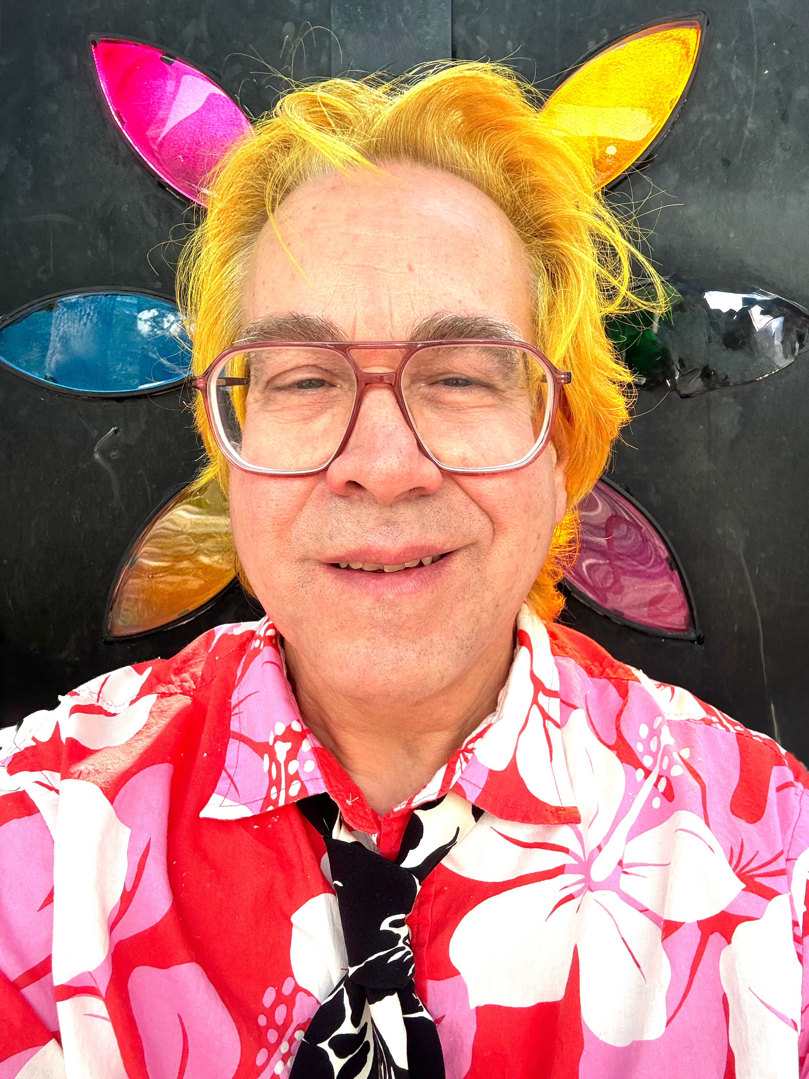 Person with colorful hair and glasses, wearing a floral shirt, in front of a black background with colorful shapes.
