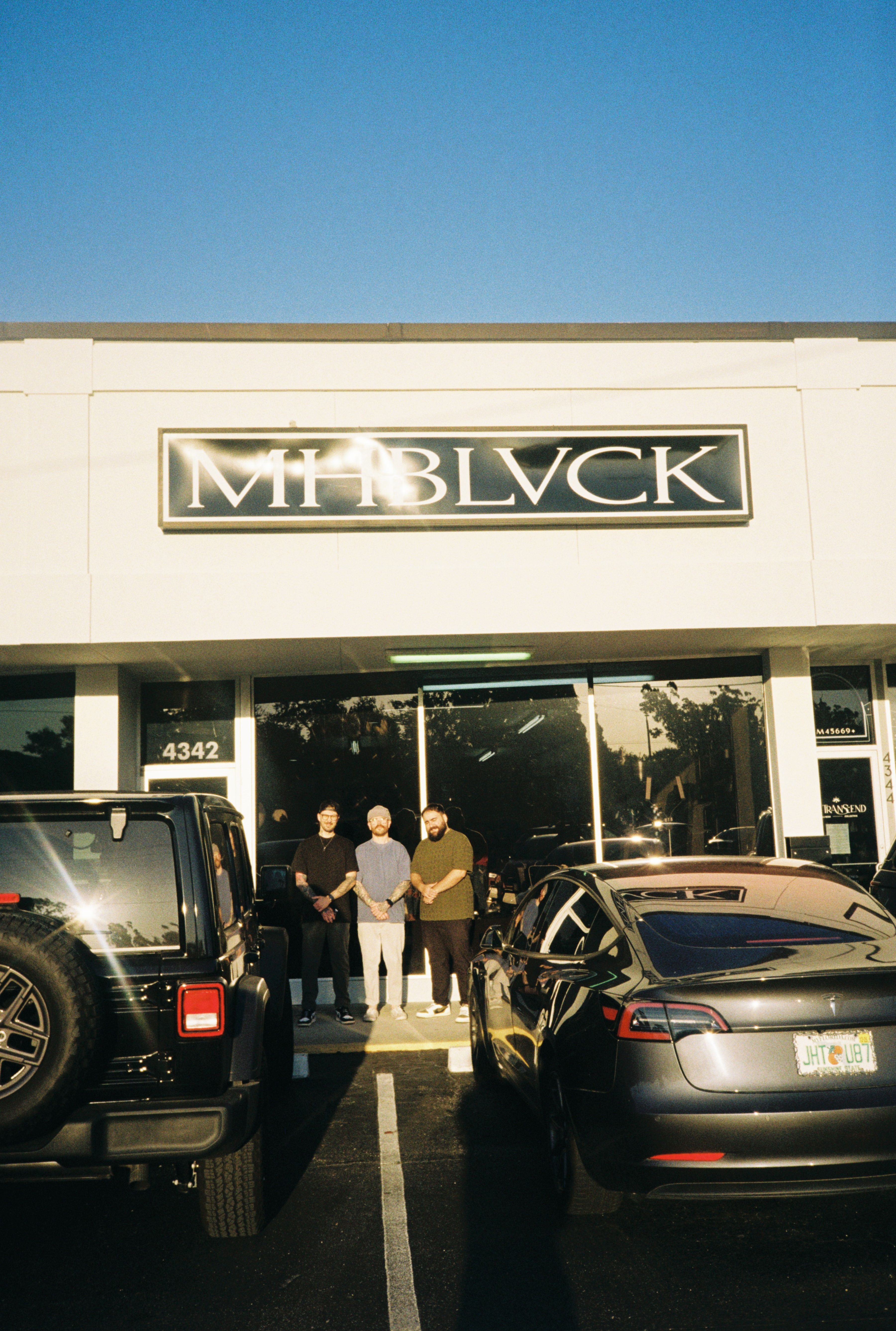 People walking outside a building with a sign reading 'The Black' and parked cars nearby.