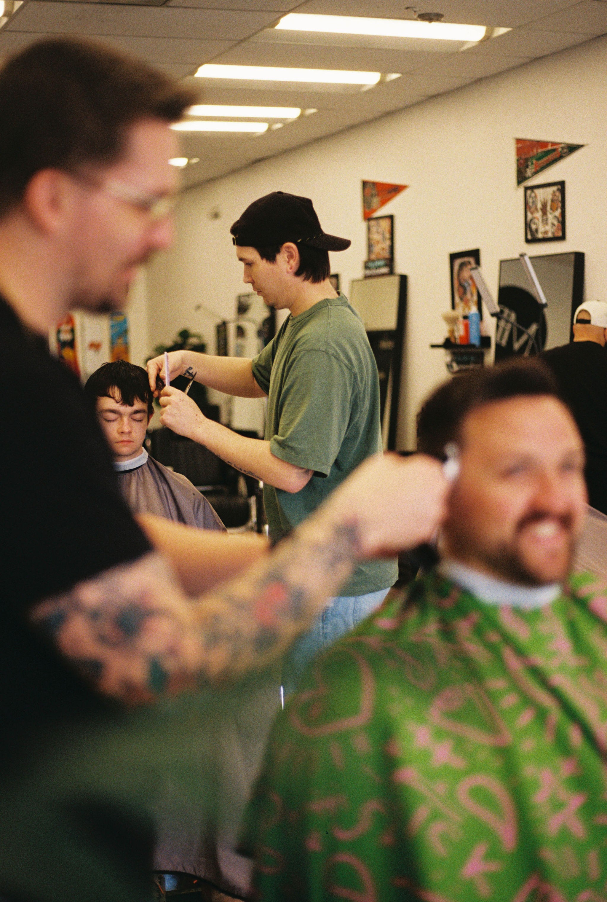 Barber cutting a man's hair in a barbershop with other customers and framed pictures on the wall.