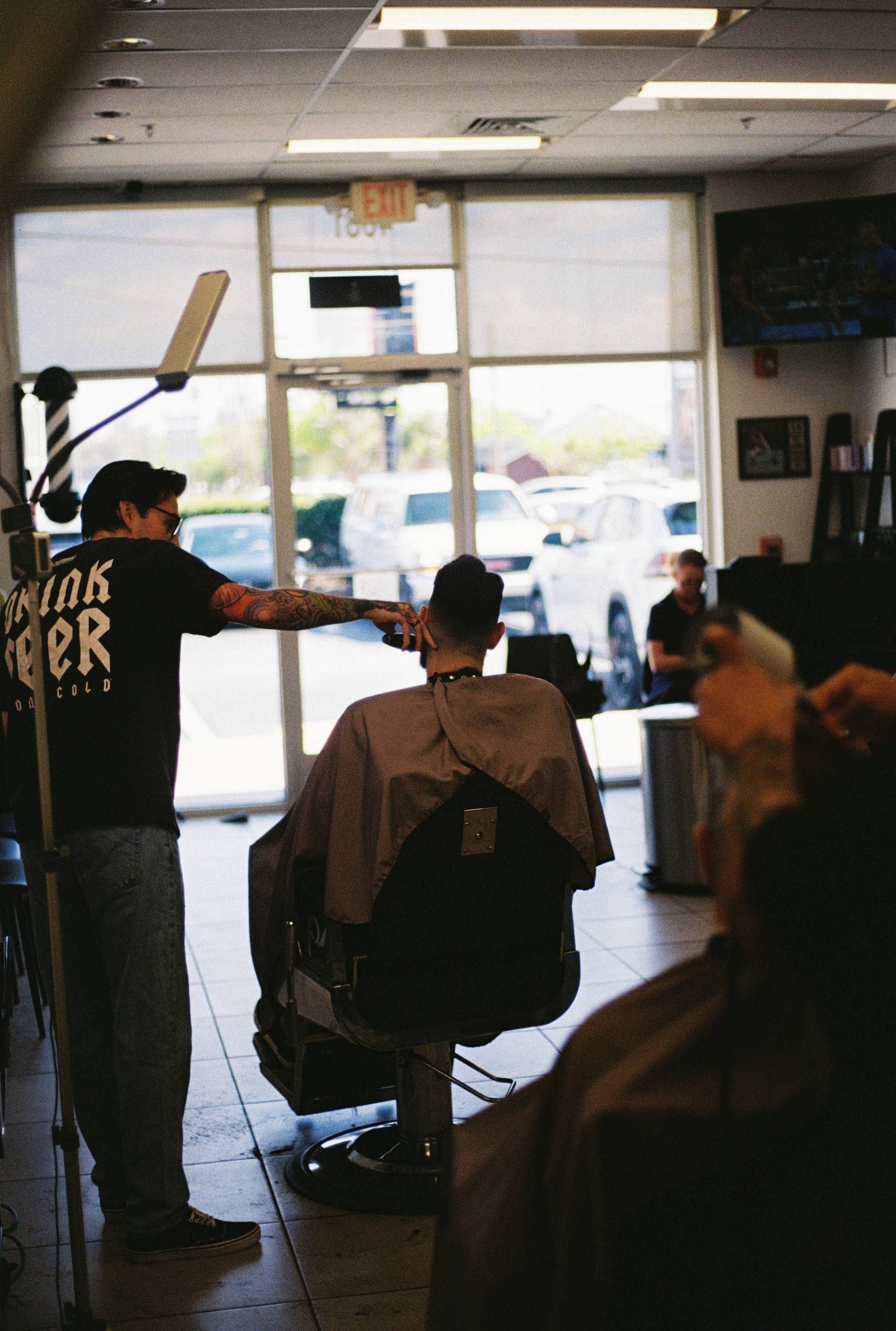 Barber cutting a person's hair in a salon with large windows and customers seated inside.