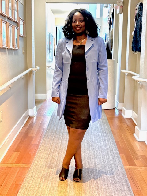 Woman standing in hallway, smiling, wearing a light blue coat over a black dress, with black heels, on a wooden floor.