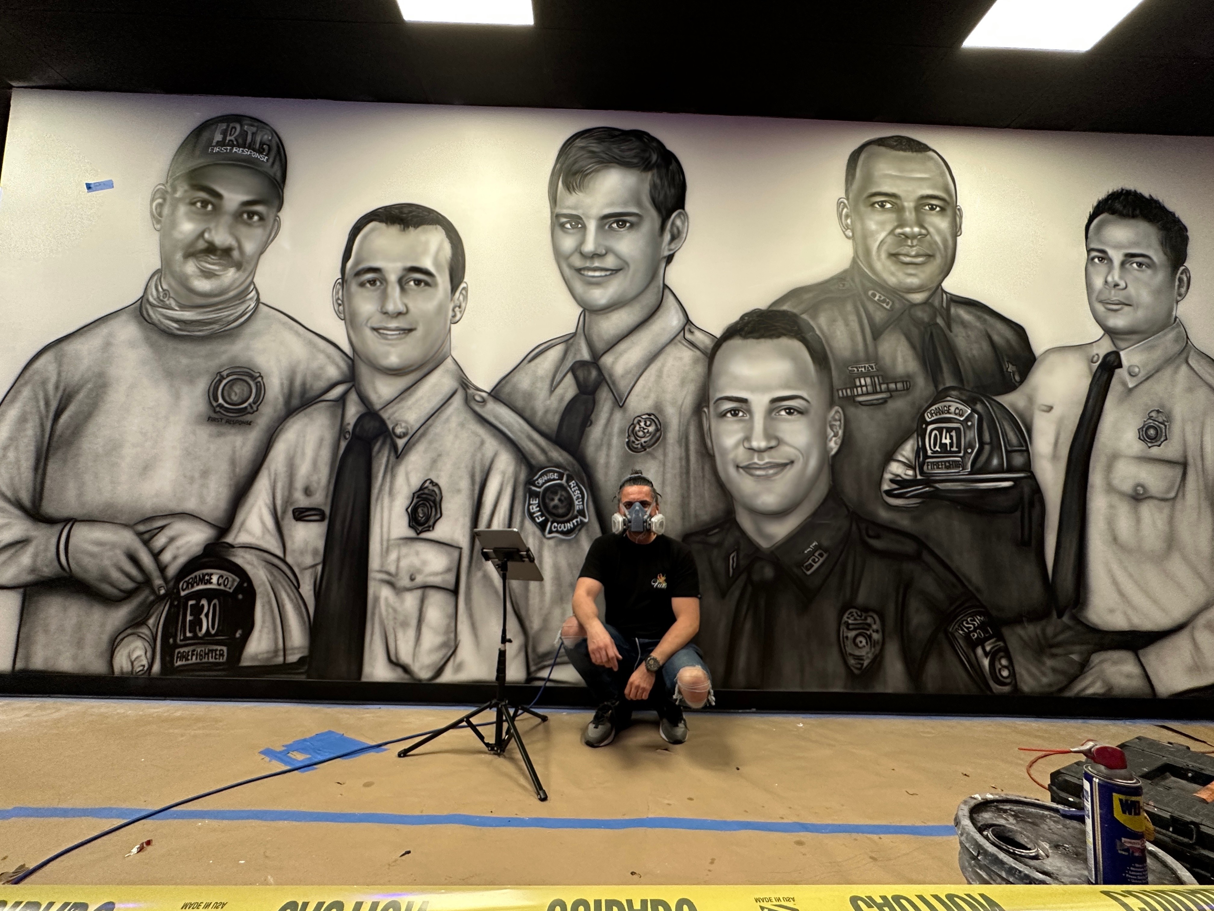 Person crouching in front of a large black-and-white mural of six police officers, with equipment on the floor nearby.