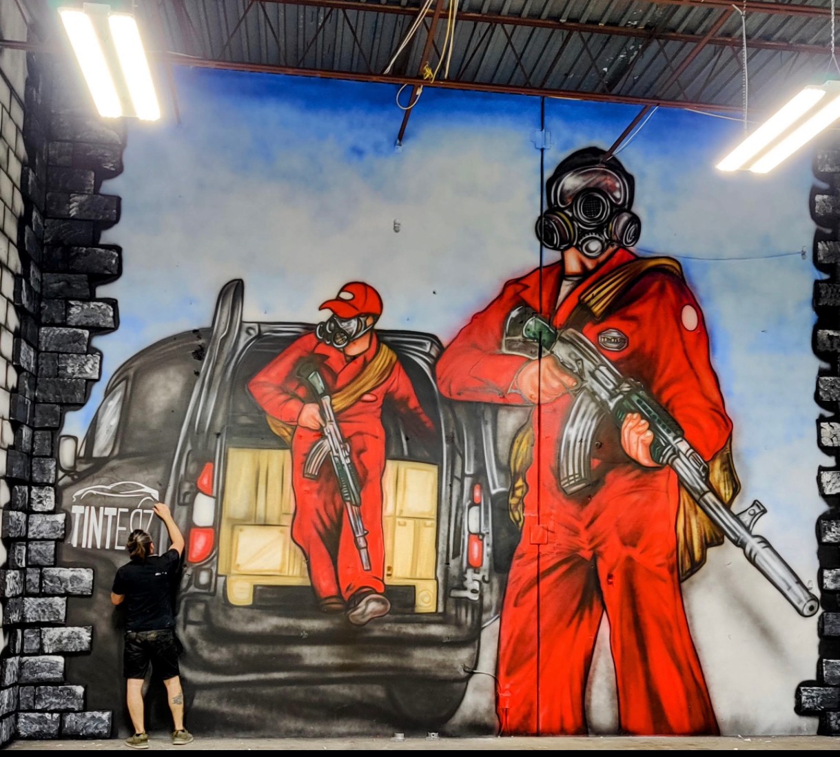Two people in red jumpsuits and gas masks stand near a black vehicle with an open trunk, graffiti background, and smoke.