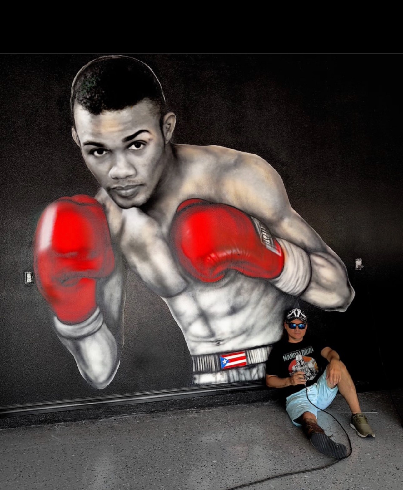Mural of a boxer with red gloves, muscular build, and intense expression, with a person sitting nearby against a dark background.