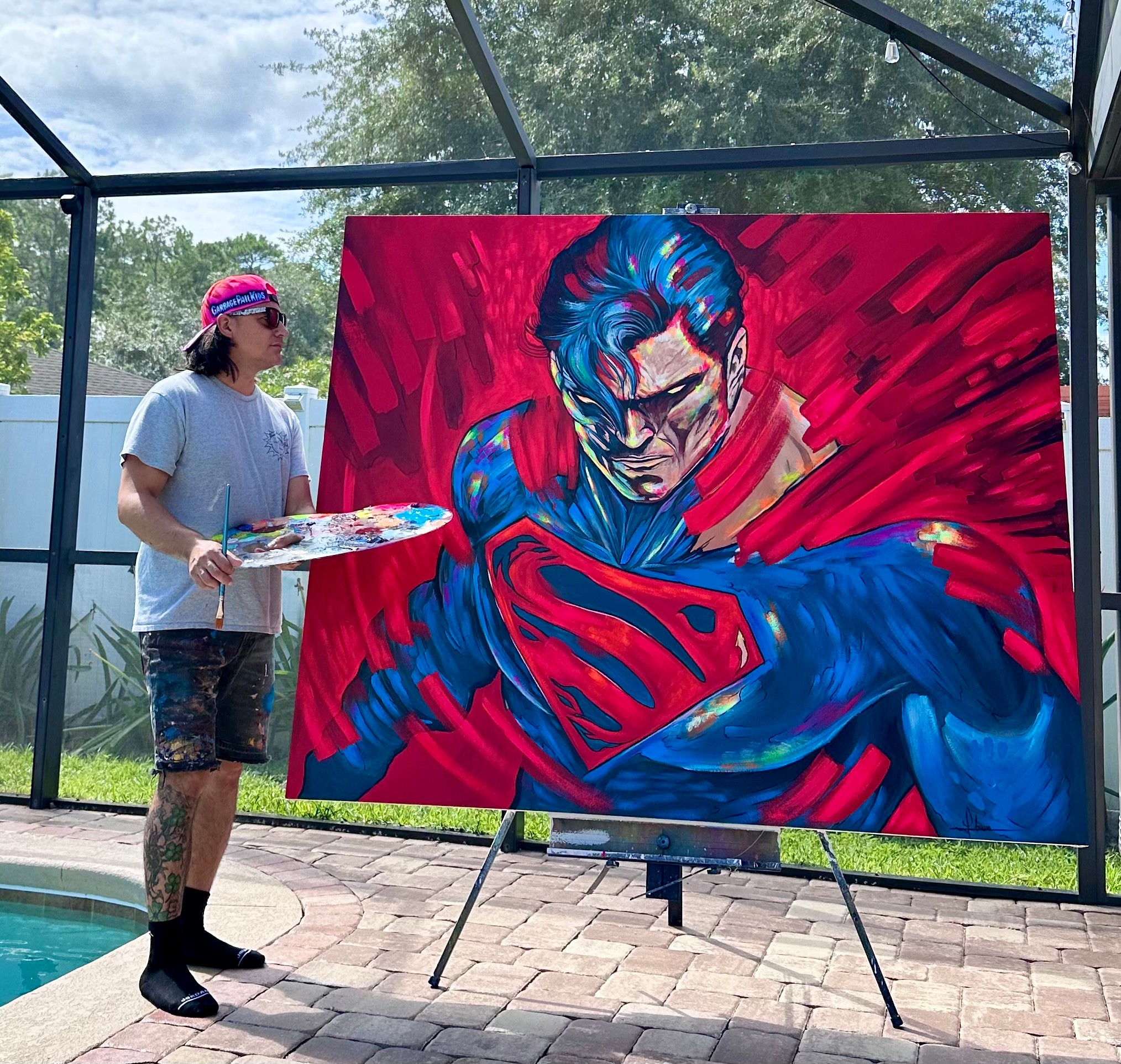 Artist standing next to a large Superman painting outdoors, with trees and sky visible in the background.