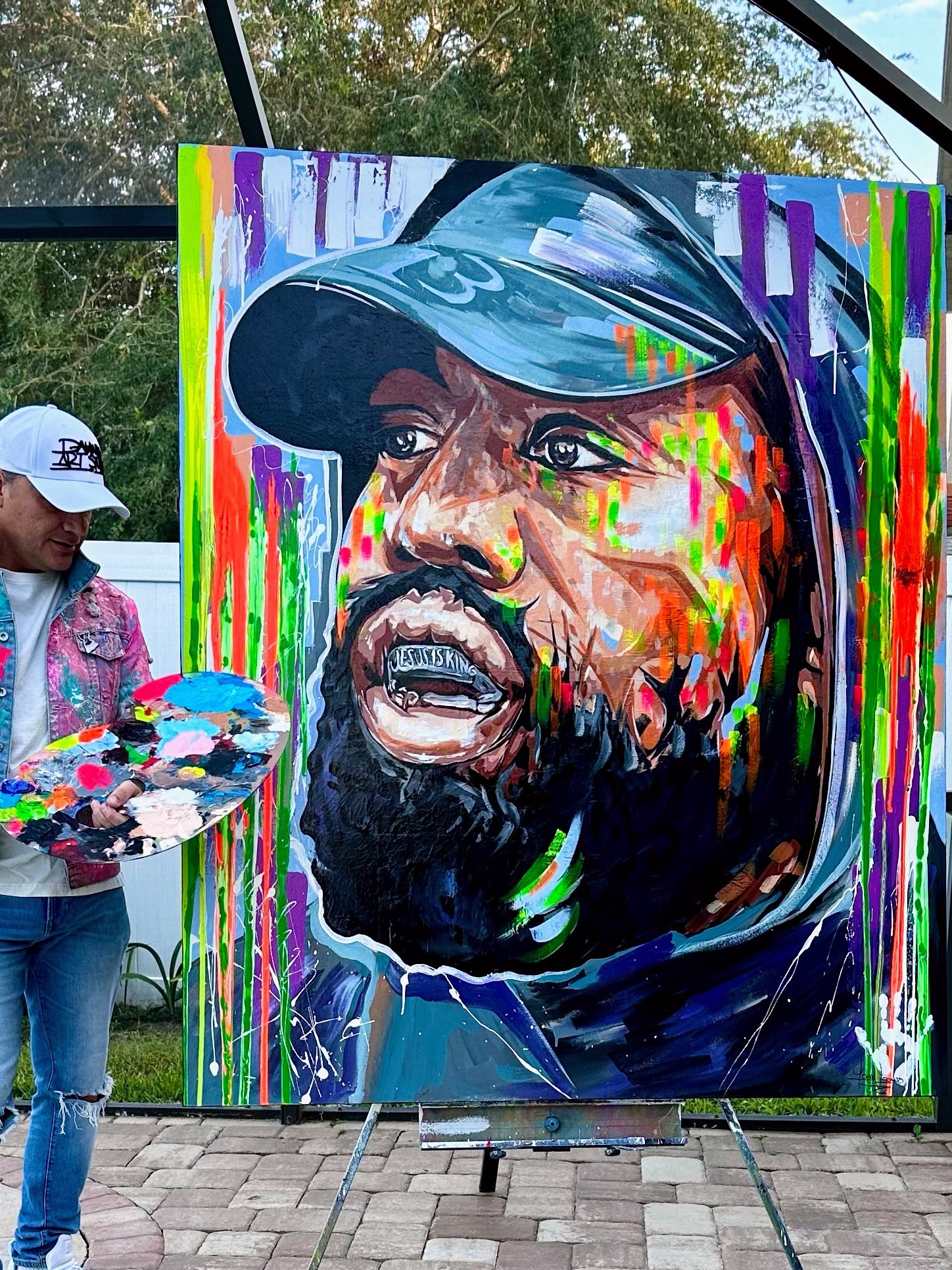 Colorful portrait of a man with a beard wearing a cap, painted on a large canvas outdoors.
