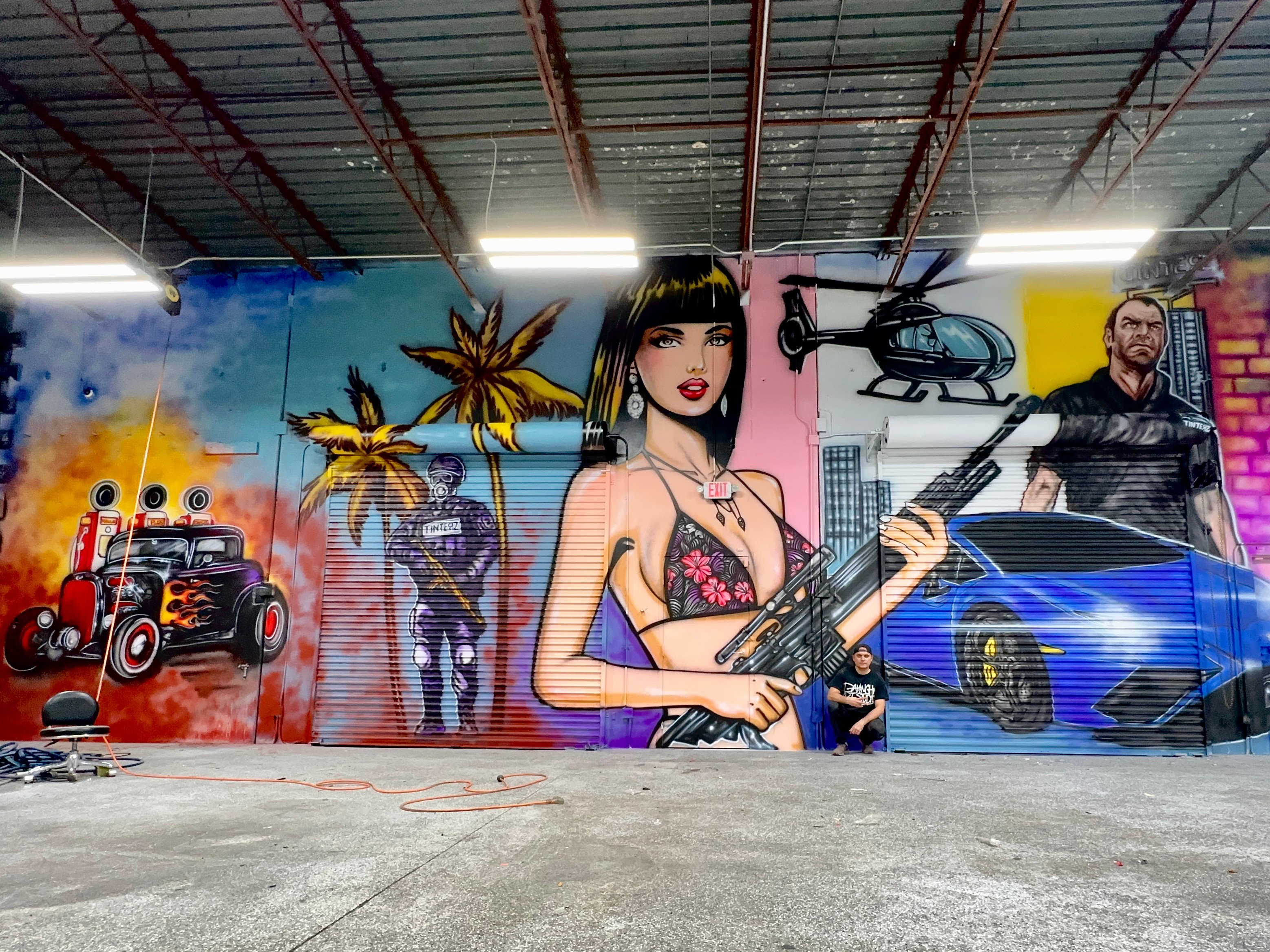 Colorful mural featuring a woman with a guitar, palm trees, a helicopter, a sports car, and a man, painted on a wall.