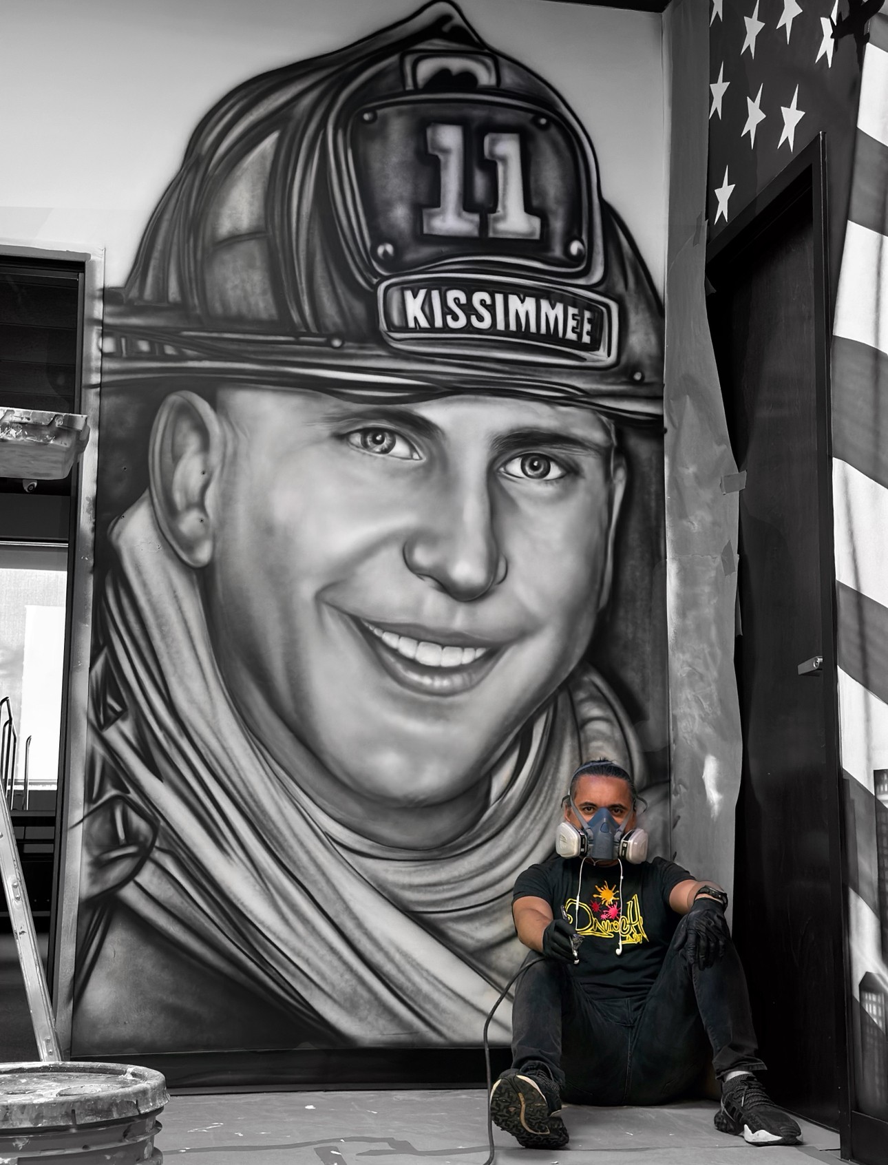 Person sitting in front of a large mural of a smiling firefighter wearing a helmet with the number 11 and the word KISSIMME, holding a camera.