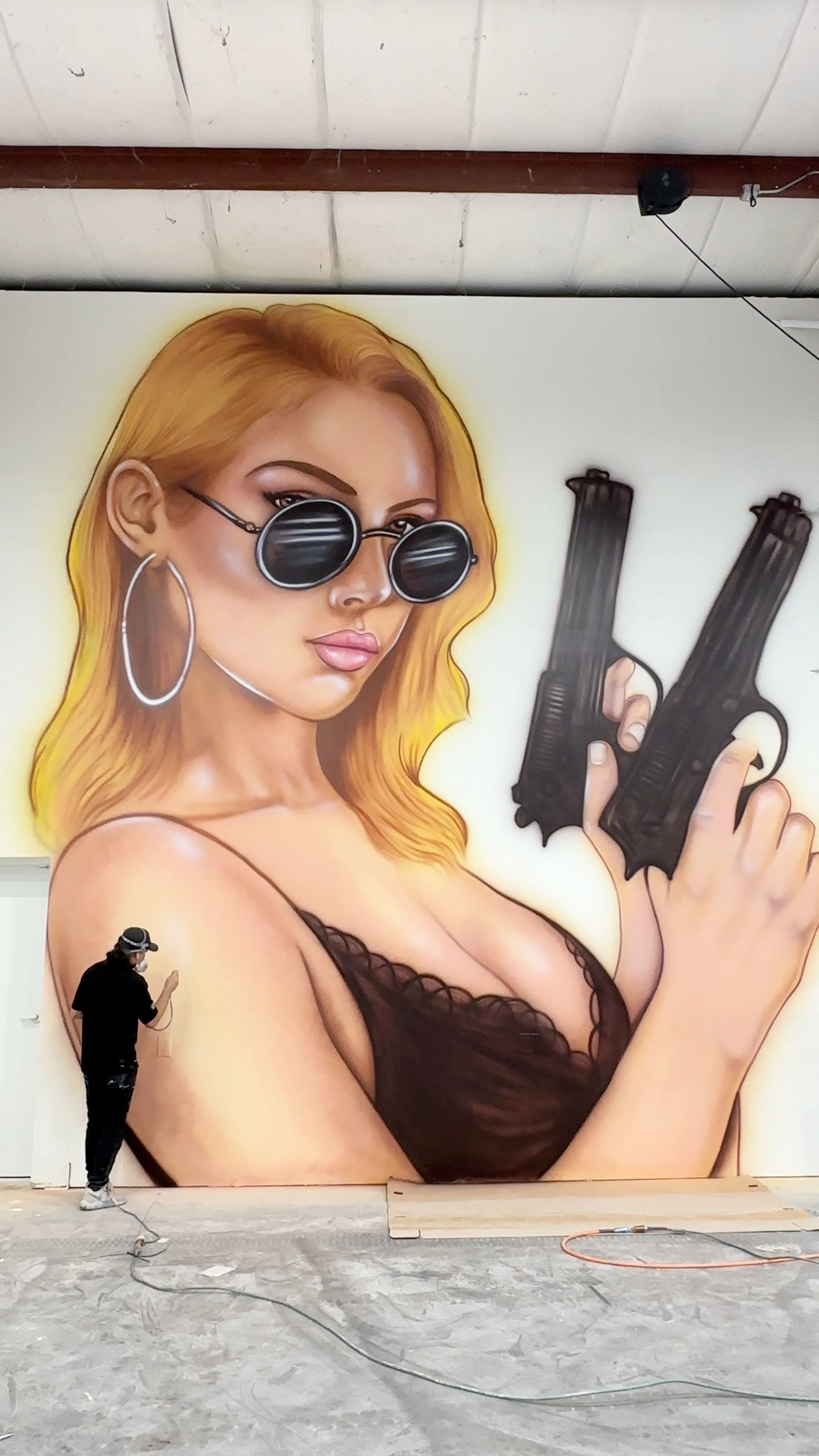 Large mural of a woman with blonde hair, sunglasses, hoop earrings, holding two guns, wearing a black top.