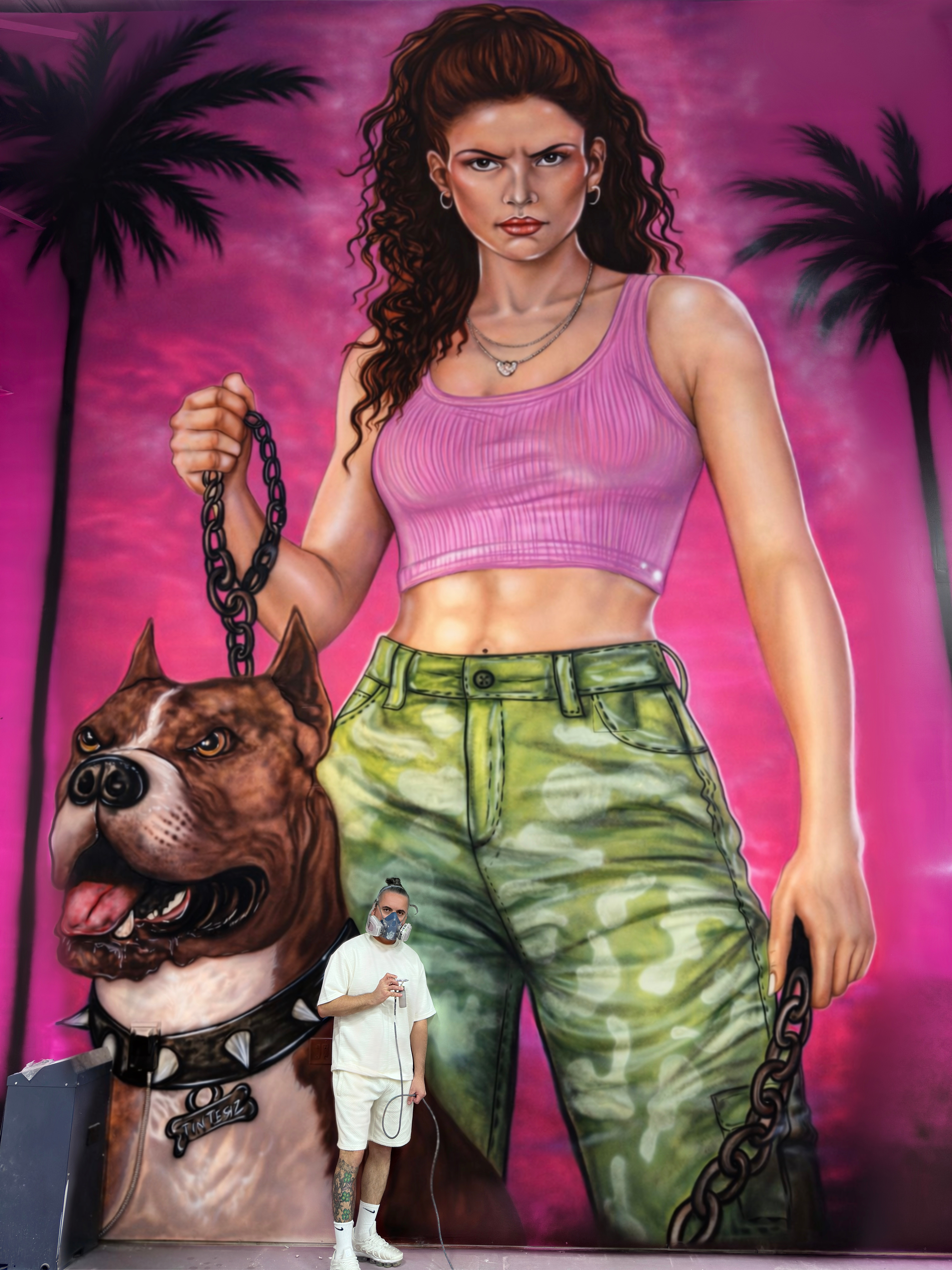 A woman with curly hair in a pink crop top and green camouflage pants holding a dog leash, standing in front of a mural with palm trees.