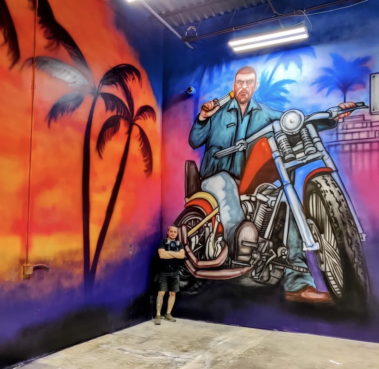Person standing in front of a colorful mural of a man on a motorcycle with palm trees and sunset background.