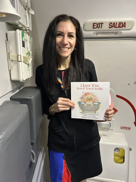 Woman with long dark hair smiling, holding a card with a cupcake illustration, standing in a room with medical equipment and an exit sign.