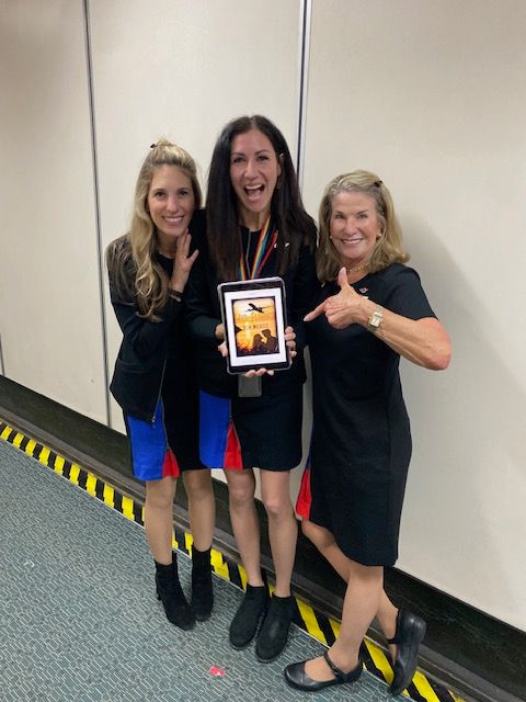 Three women standing in an elevator, one holding a tablet, all smiling and making peace signs.