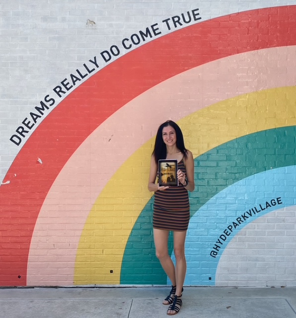 Woman standing in front of a colorful rainbow mural holding a tablet, with text 'DREAMS REALLY DO COME TRUE' and '@HYDEPARKVILLAGE'.