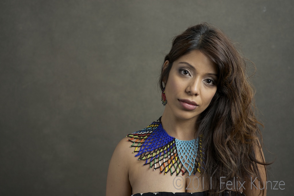 Young woman with long, wavy hair wearing a colorful, beaded necklace and earrings, against a plain background.