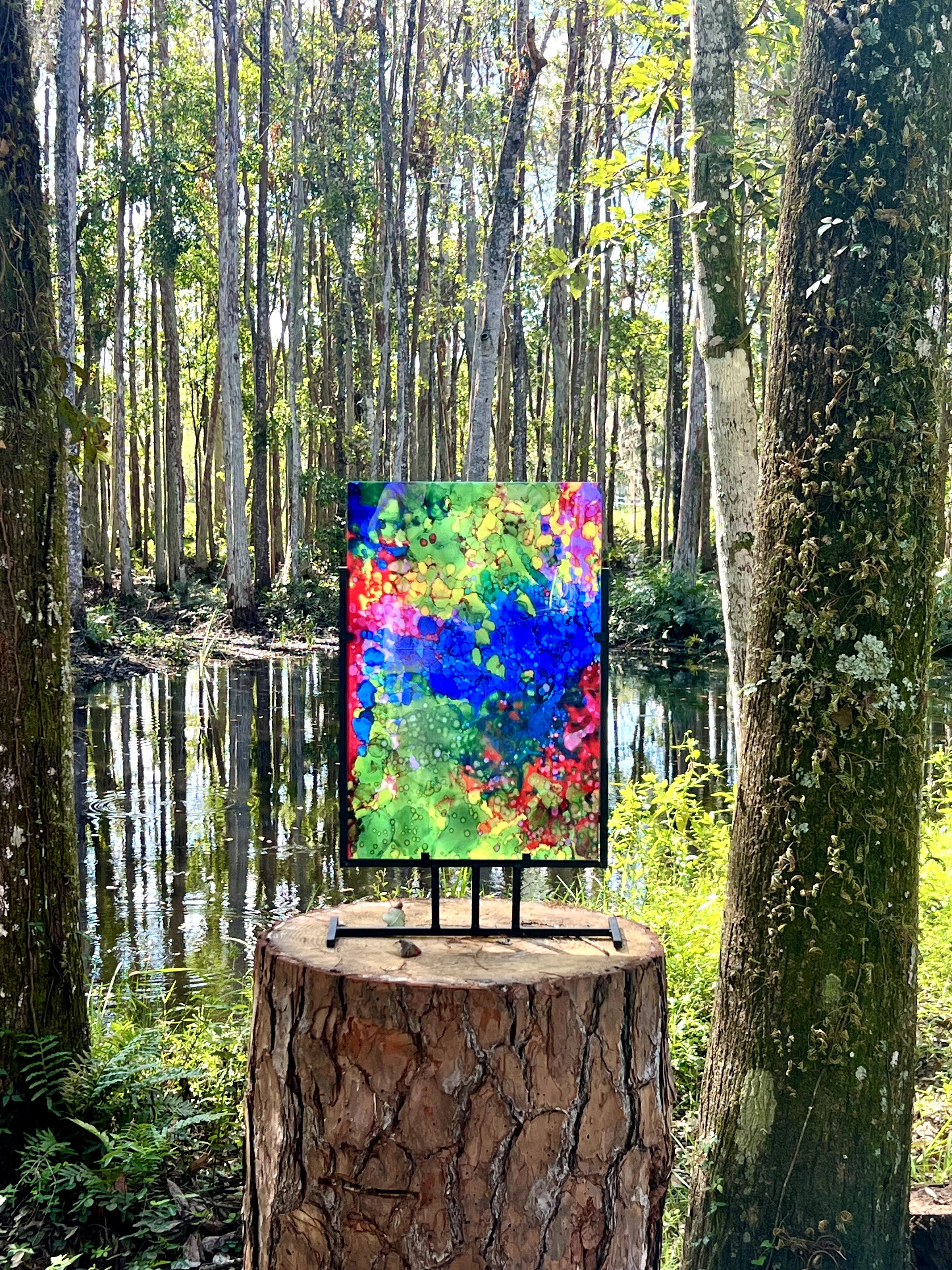 Colorful abstract painting on an easel placed on a tree stump in a forest with tall trees and a water body.