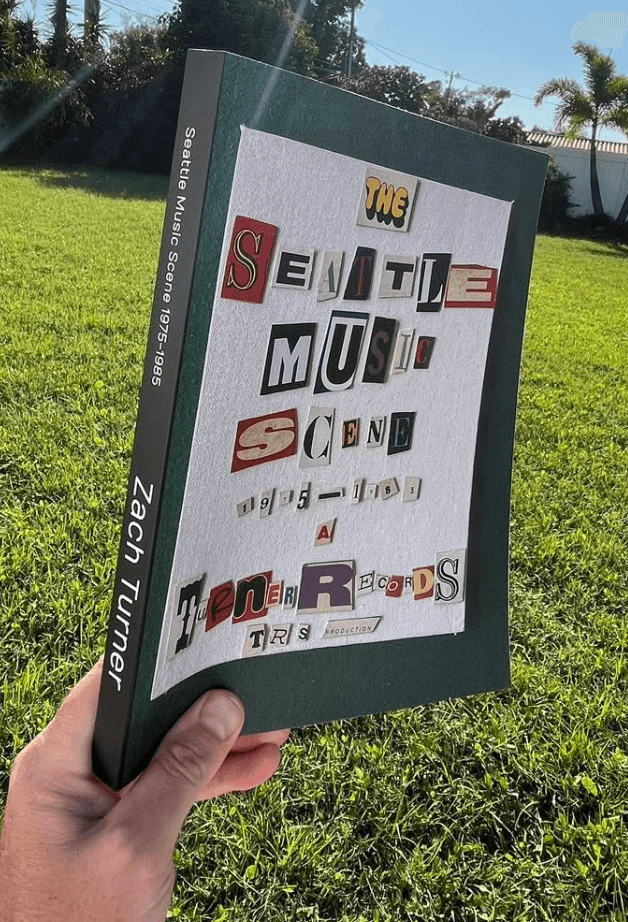 Person holding a book outdoors with a grassy background and trees, the book cover has cut-out letters and words.