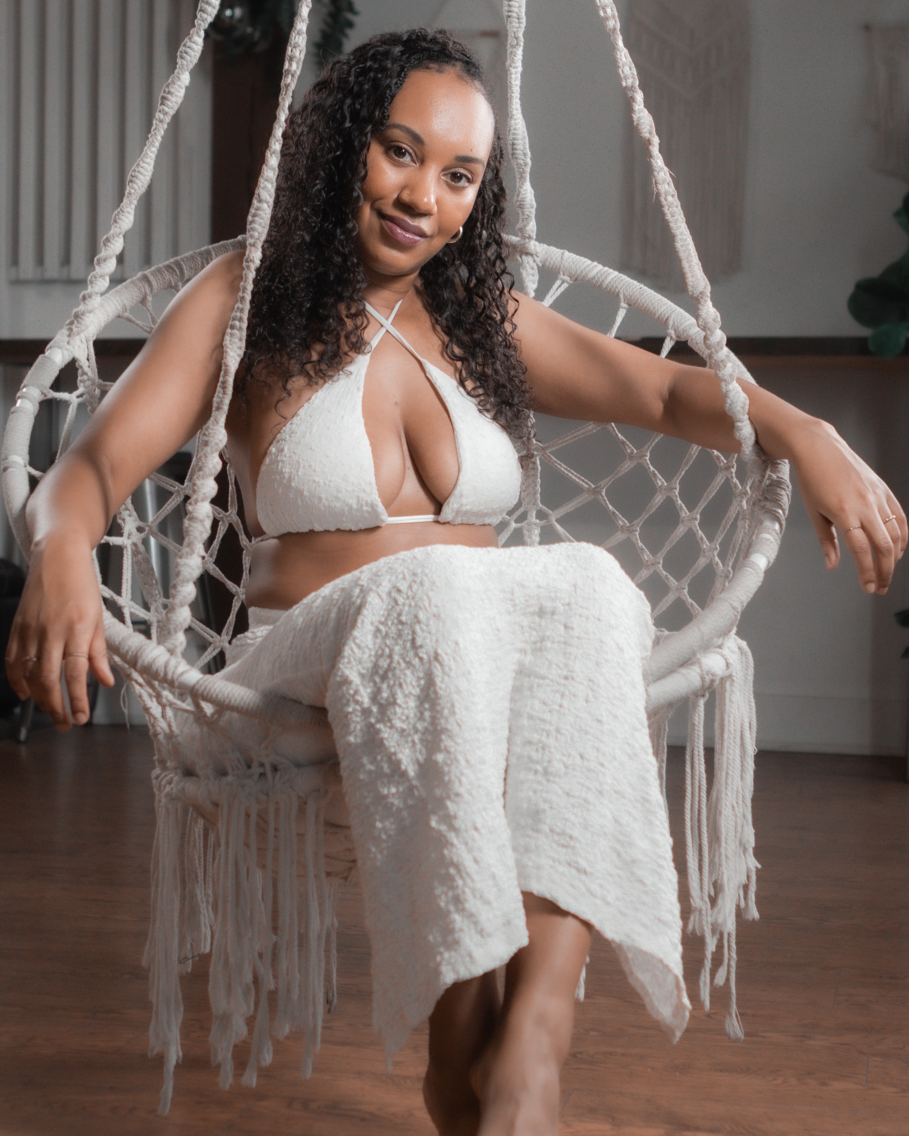Woman sitting on a hanging chair, wearing a white outfit, smiling, with a wooden floor and indoor background.