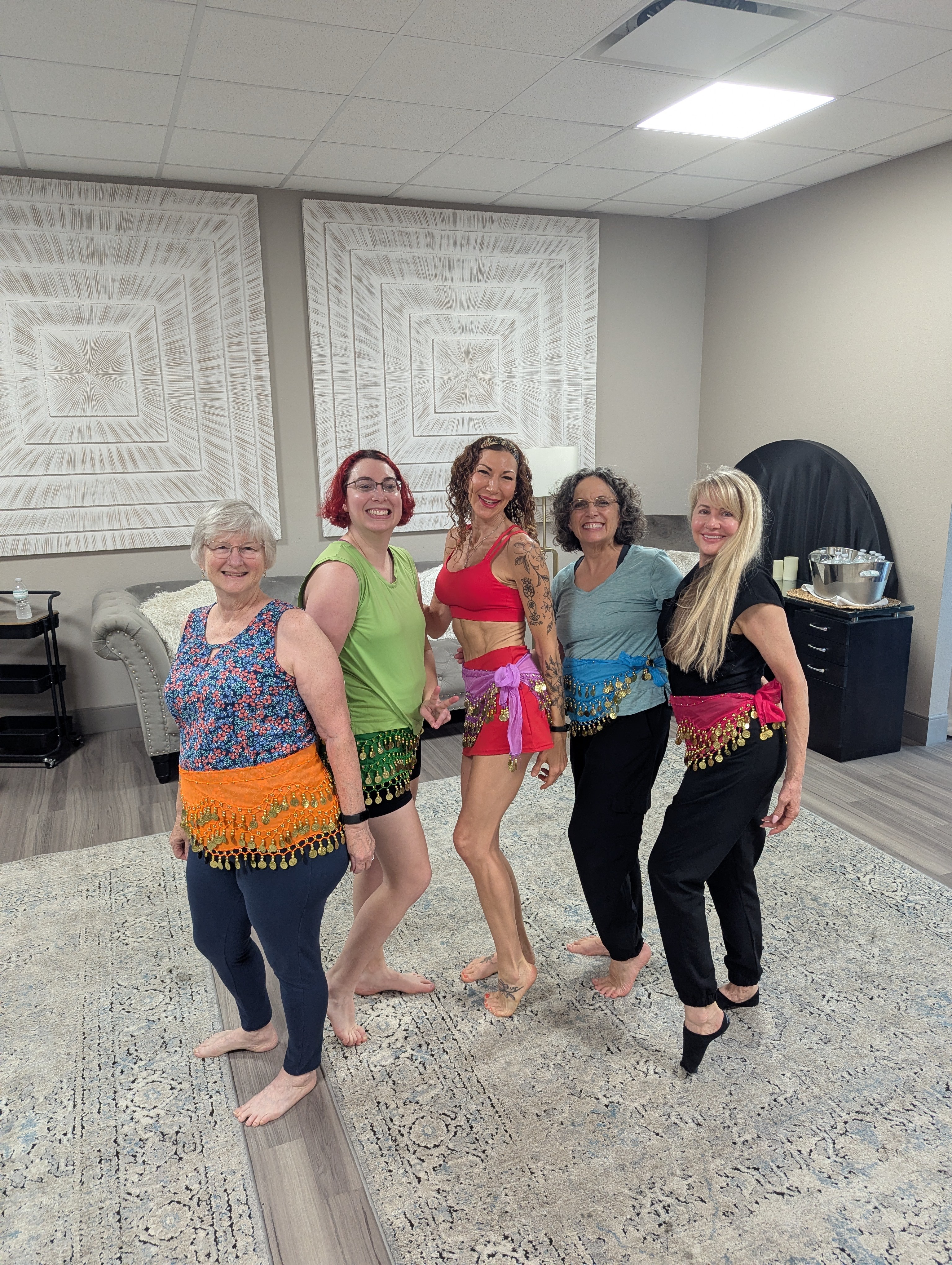 Five women standing together indoors, smiling, with patterned curtains and a carpeted floor in the background.