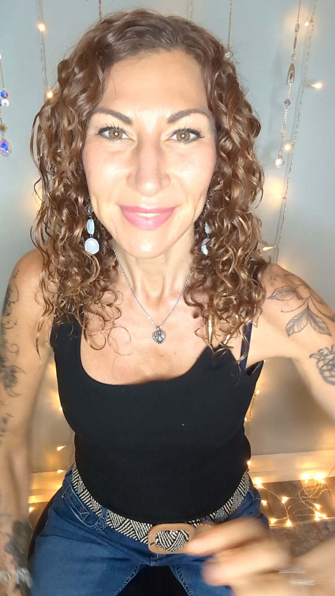 Woman with curly hair smiling, wearing a black top and jewelry, standing indoors with string lights in background.