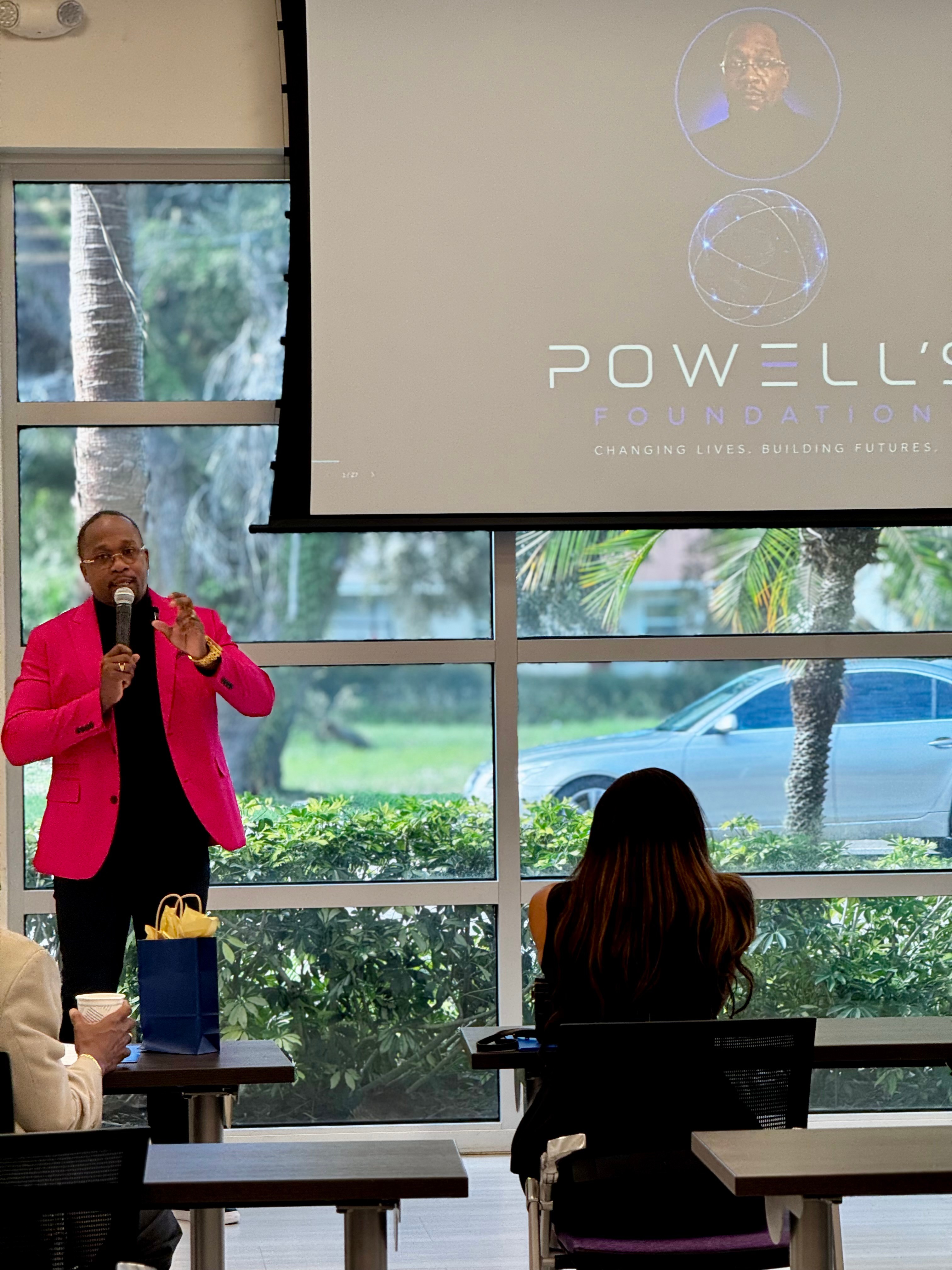 Person in pink blazer speaking into microphone in front of large window with greenery outside.