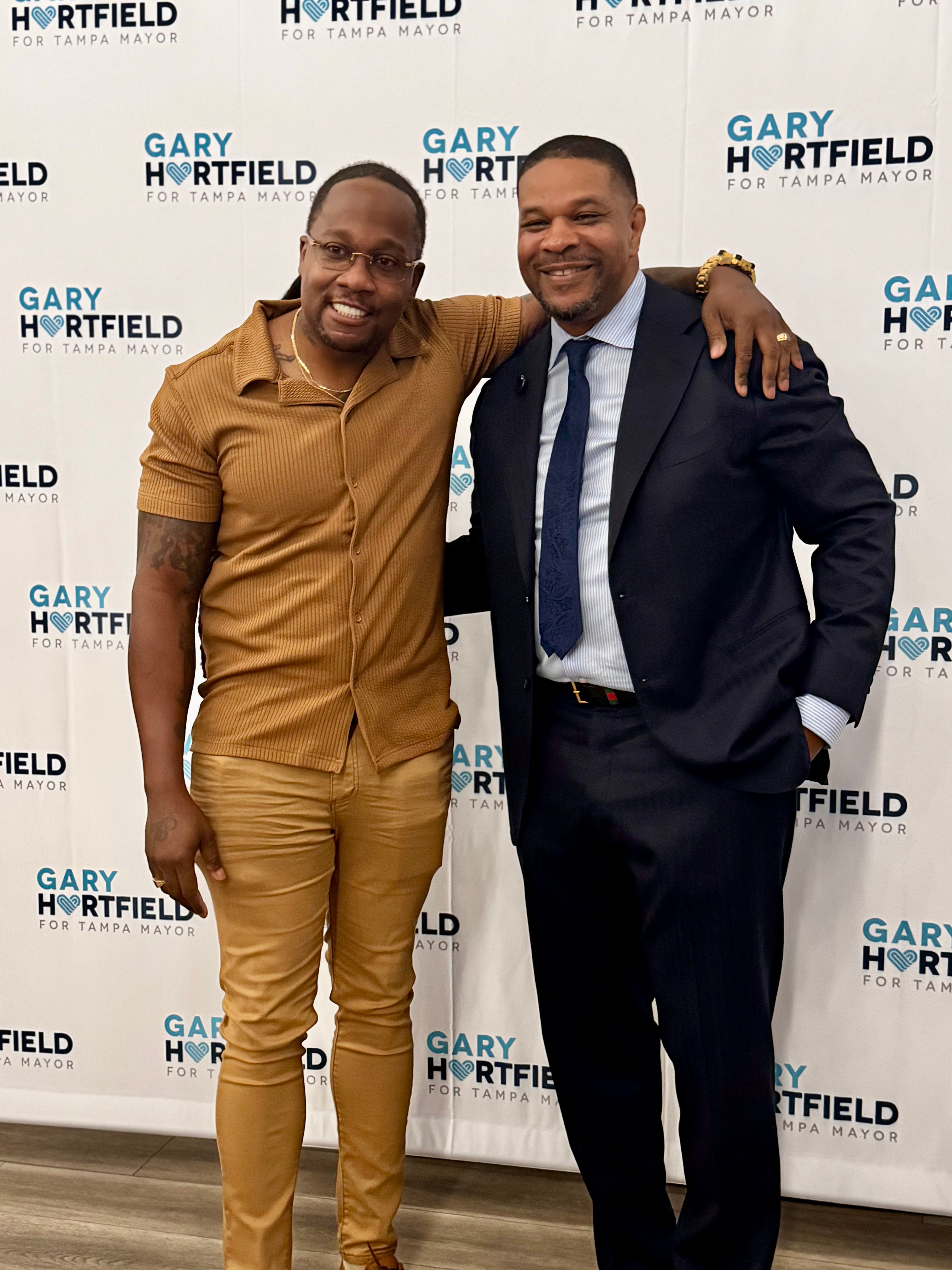 Two men smiling and standing close, one with arm around the other, in front of a campaign backdrop.