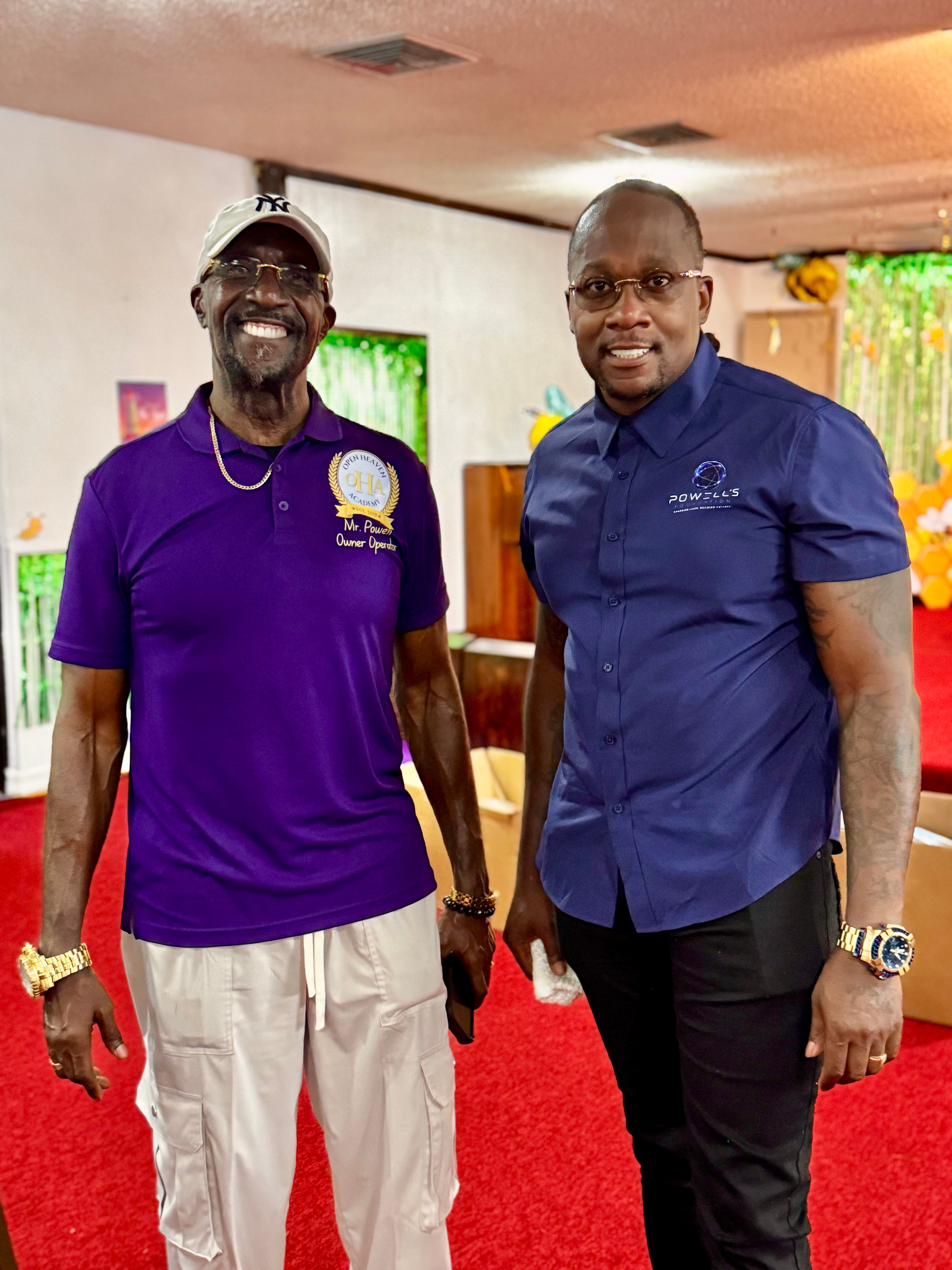 Two men standing indoors, smiling, one in a purple shirt and the other in a blue shirt, with a red carpet and colorful background.