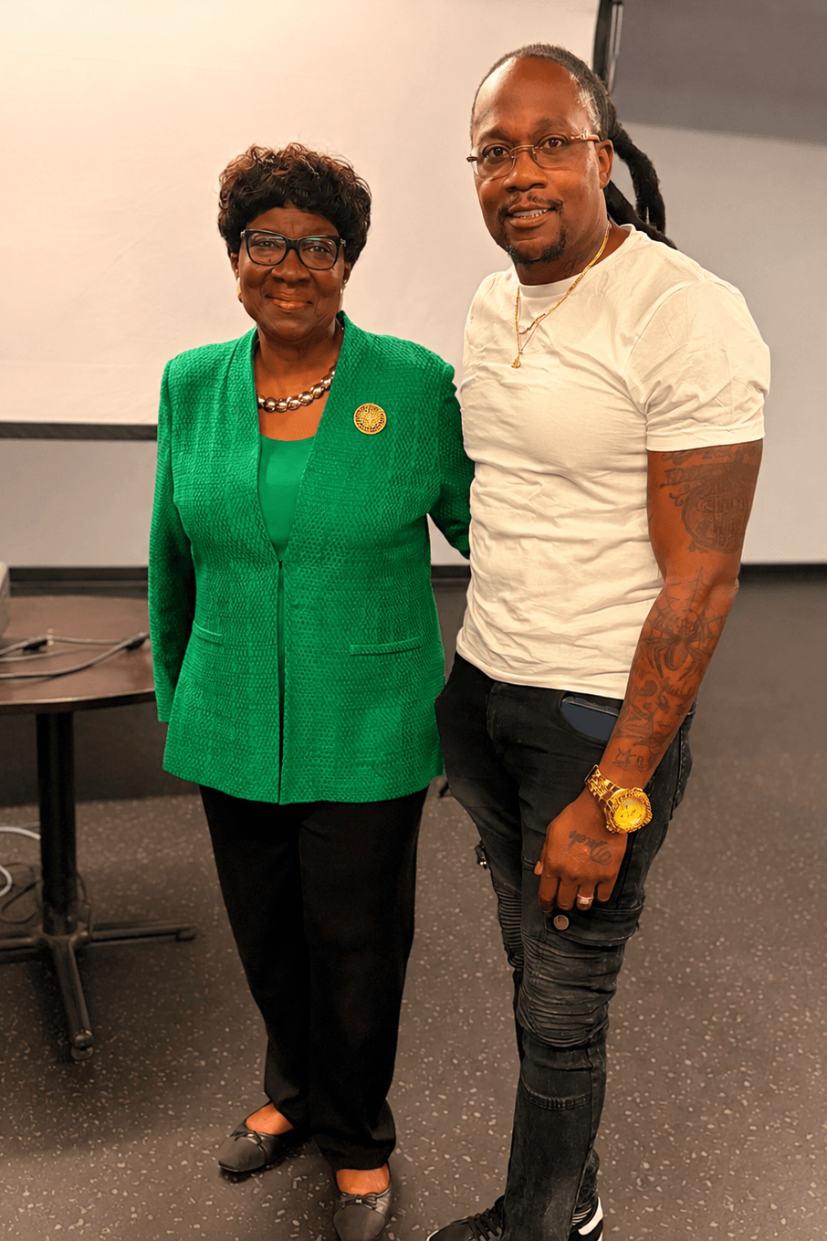 A woman in a green blazer and a man in a white t-shirt stand together indoors, smiling at the camera.