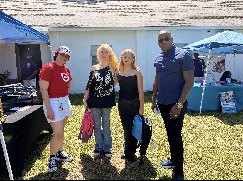 Four people standing outdoors in front of a building, with tents and tables nearby, on a sunny day.