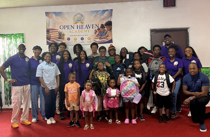 Group of children and adults in a room with a banner reading 'Open Heaven Academy' on the wall.