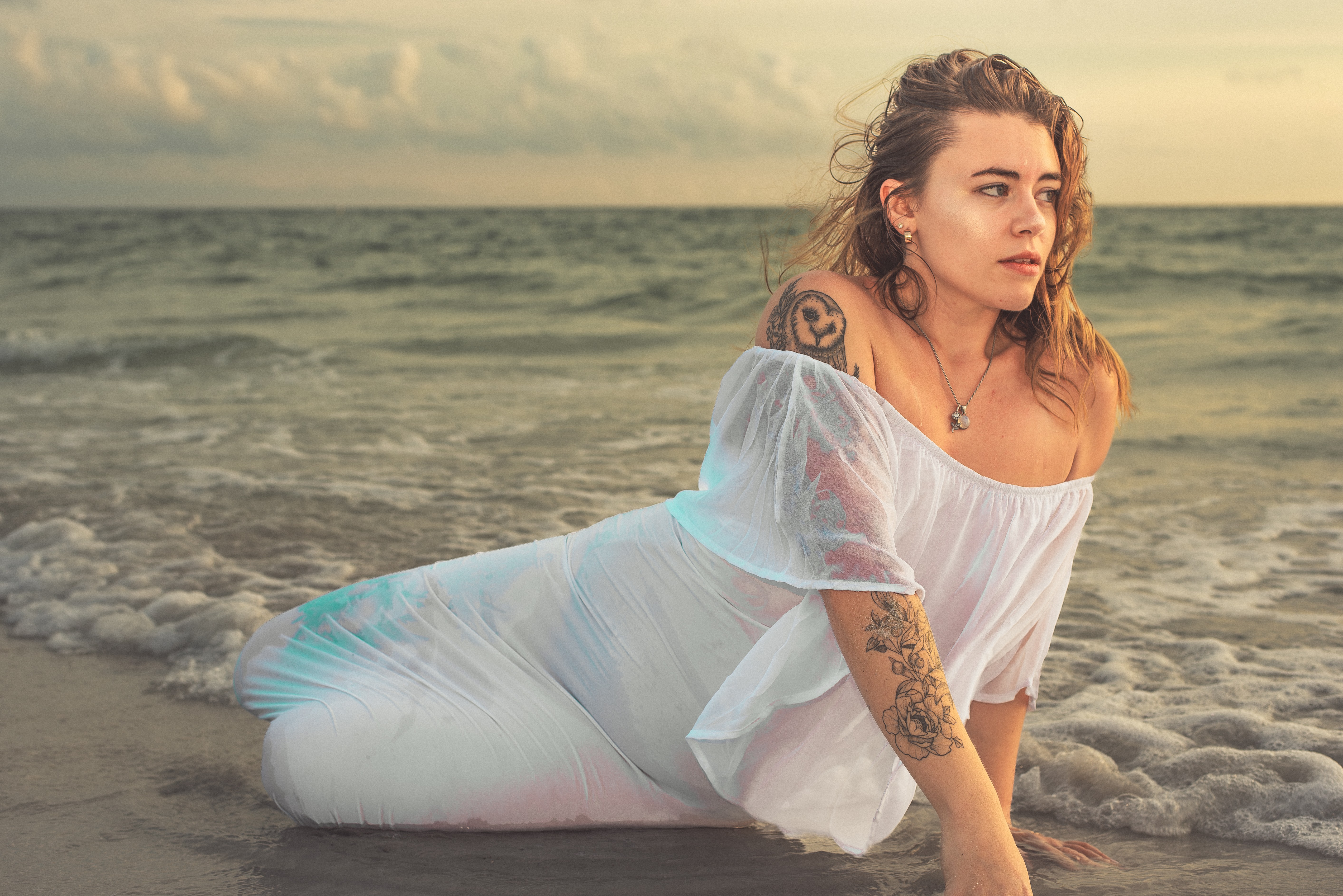 Woman sitting on the beach with ocean waves in the background, wearing a white off-shoulder dress, looking to the side.