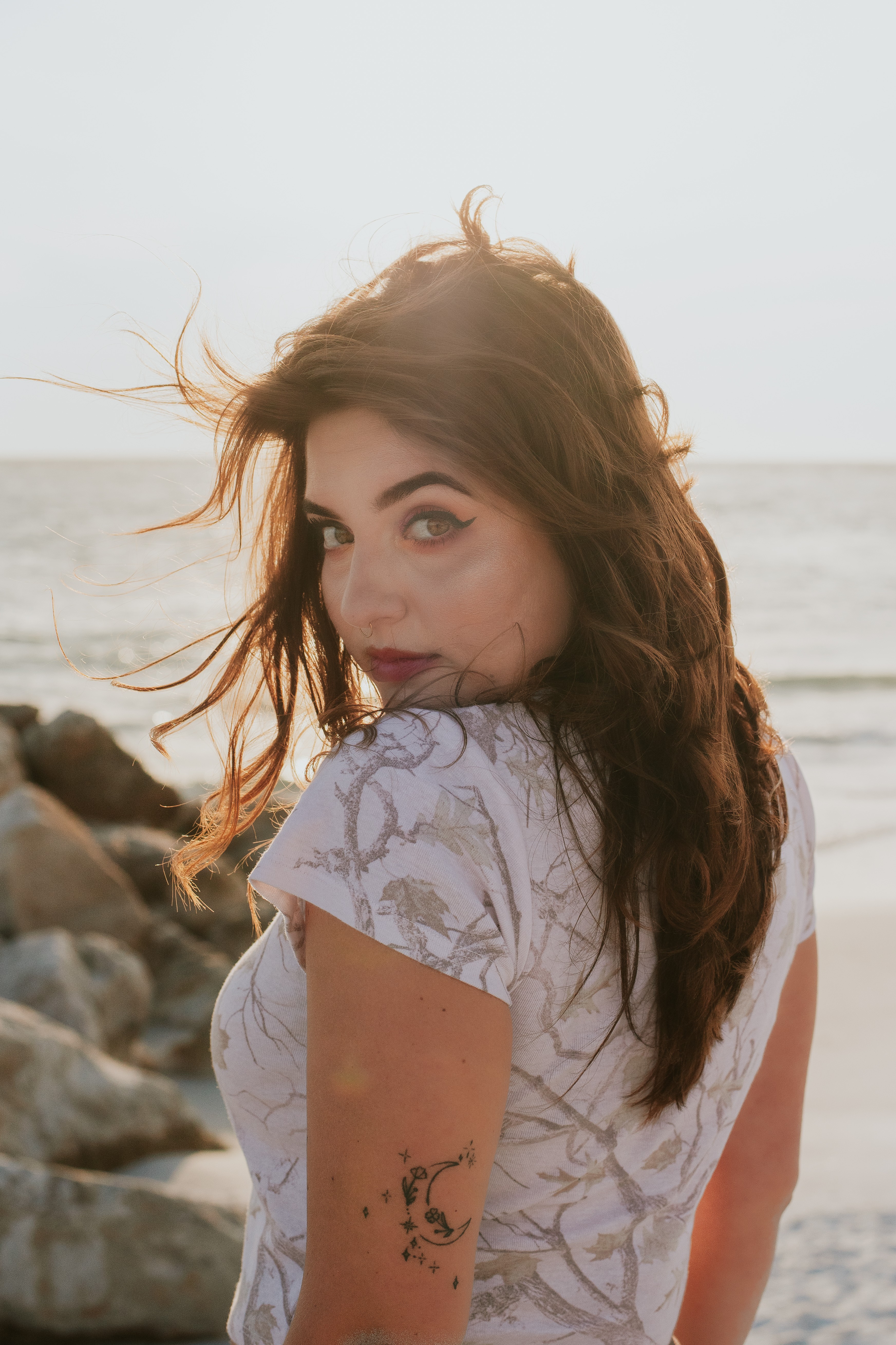 Young woman with long hair looking over shoulder at the beach, wind blowing her hair, rocks in background.