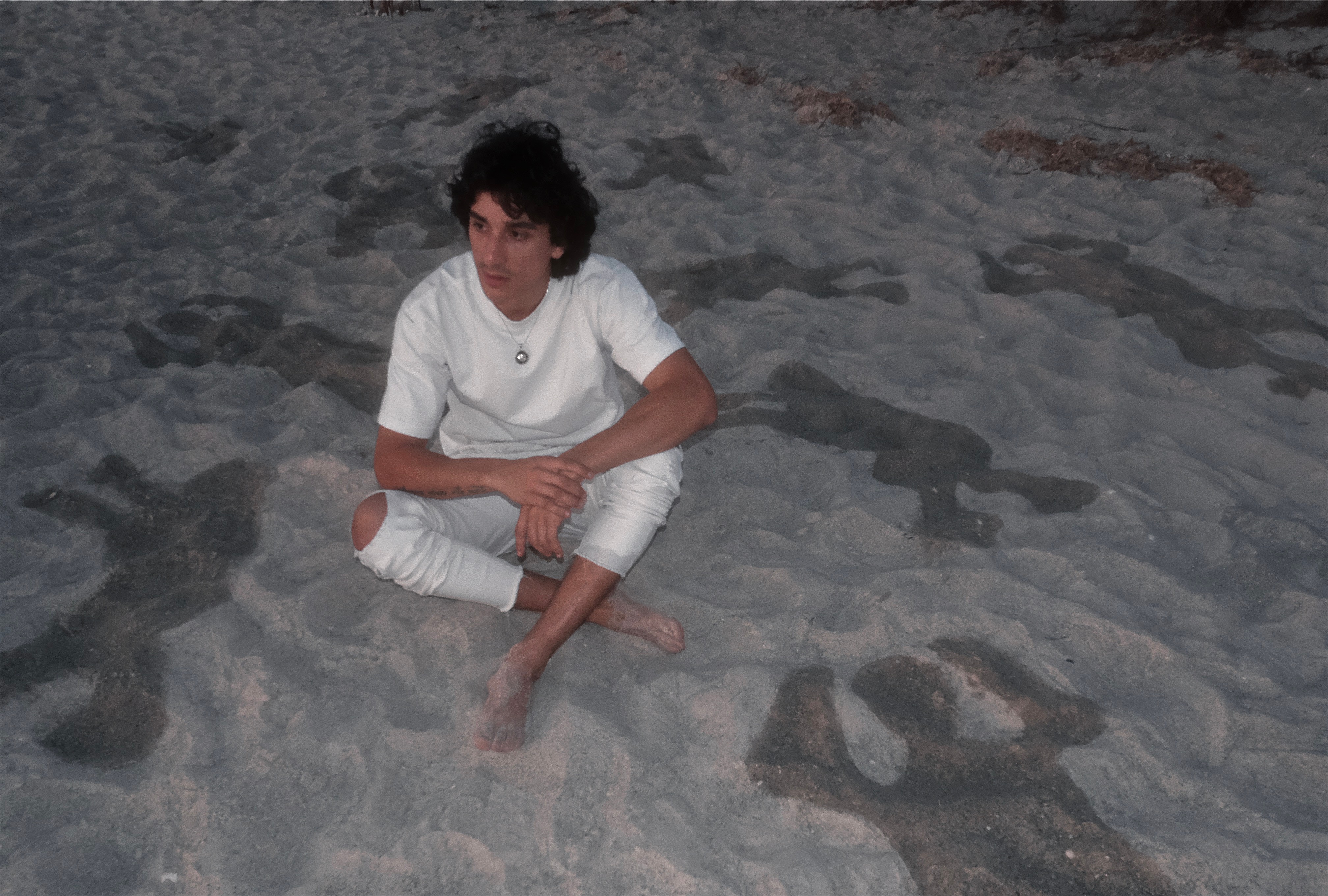Person sitting on sandy beach with rocks, wearing white clothes, looking to the side.