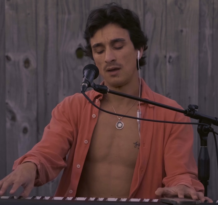 Man with dark hair singing into a microphone while playing a keyboard, wearing an open shirt and necklace, against a wooden background.