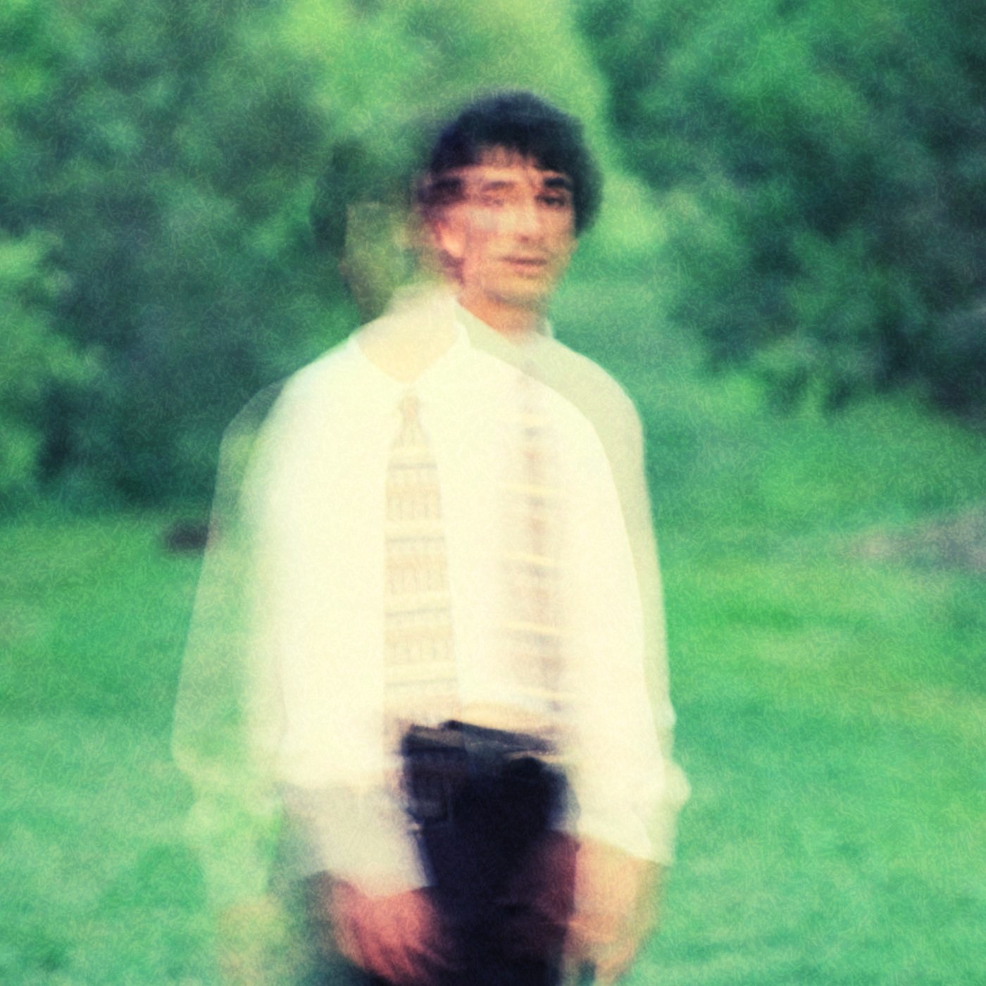 Blurry image of a person standing outdoors with green trees and grass in the background.