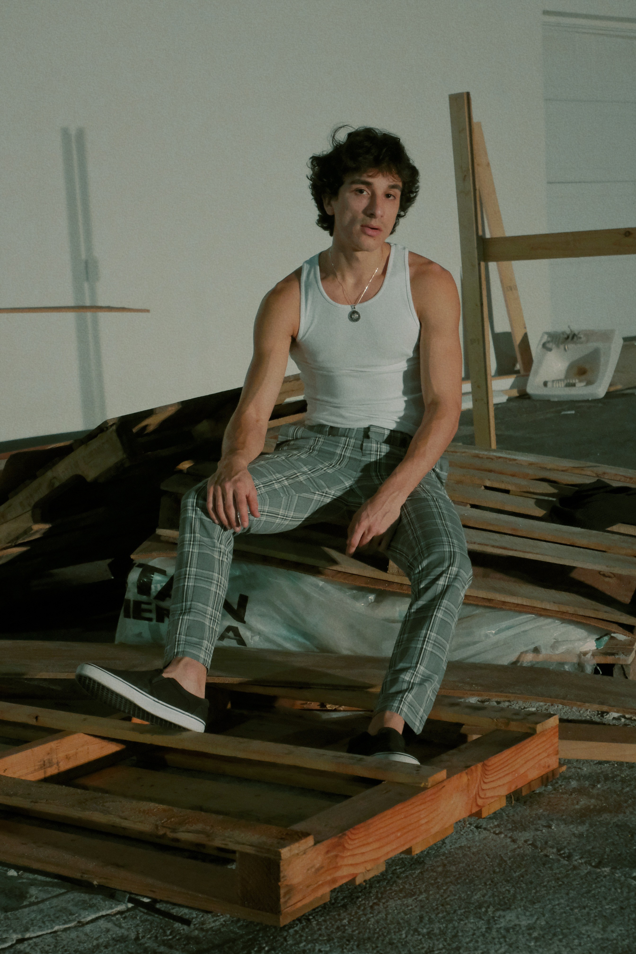 Person with curly hair sitting on a pile of wood and cardboard, wearing a white tank top and plaid pants.