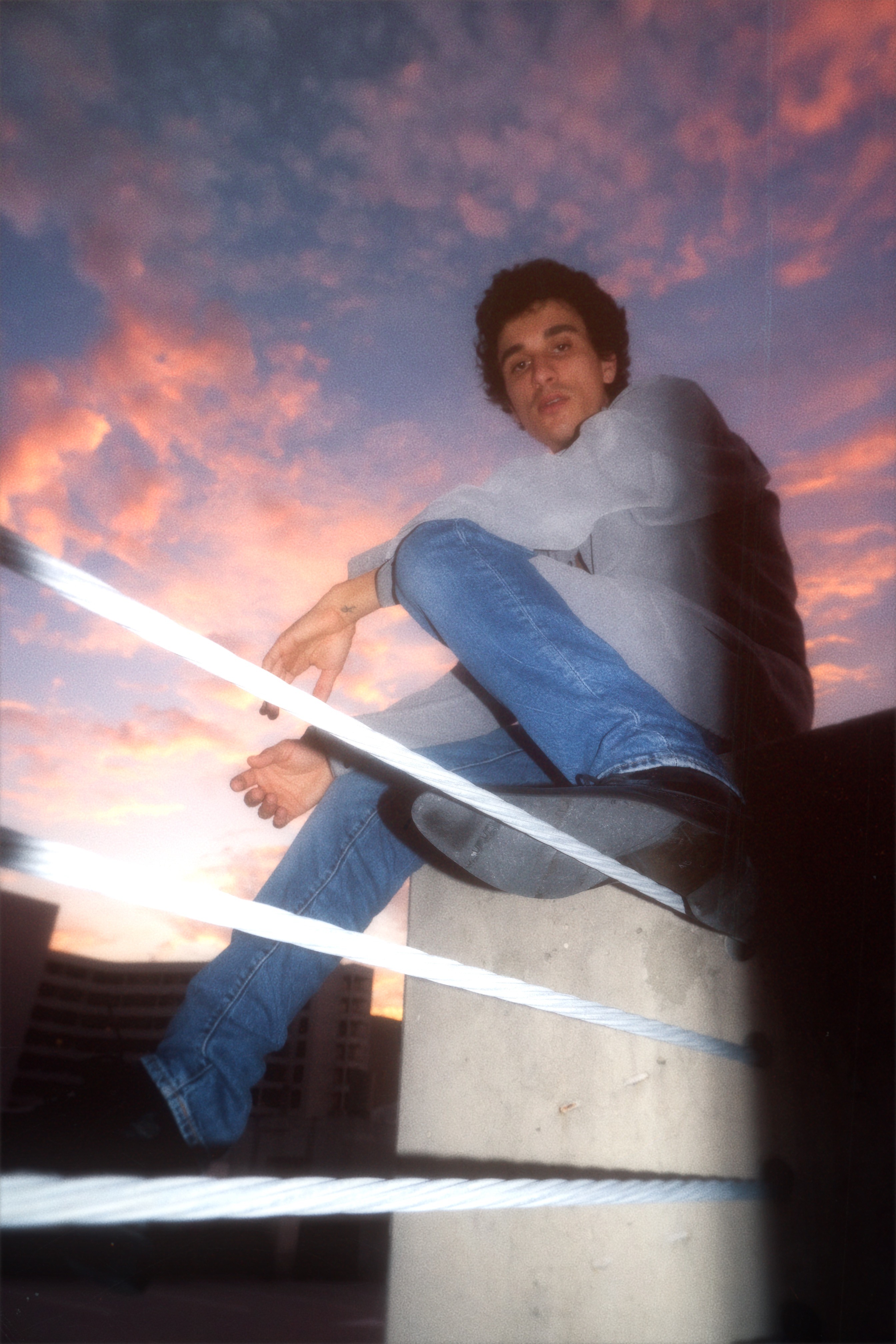 Person sitting on a concrete structure against a colorful sunset sky, smiling, wearing a gray jacket and blue jeans.