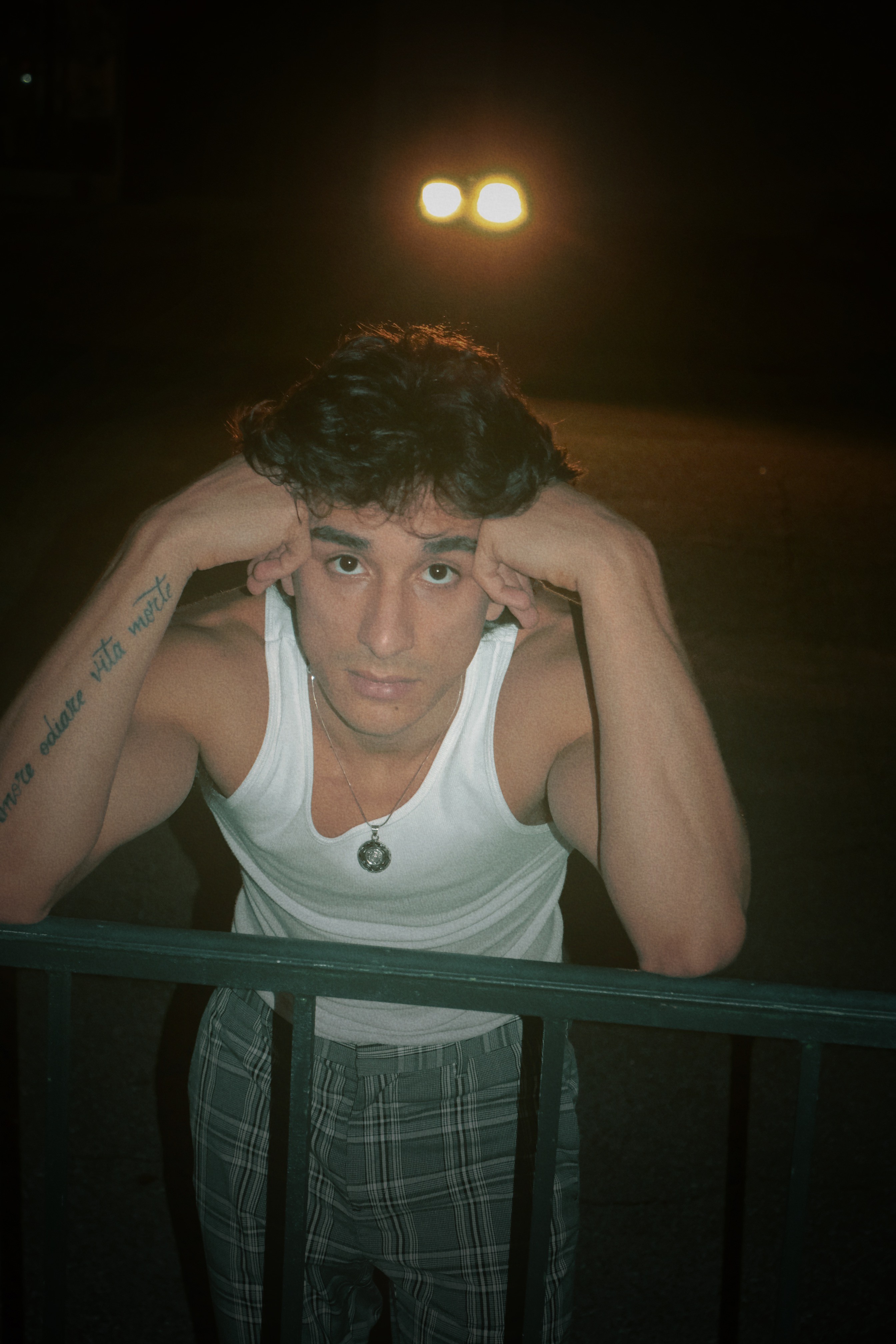 Person with curly hair wearing a white tank top and plaid pants, resting head on hands, looking at camera.